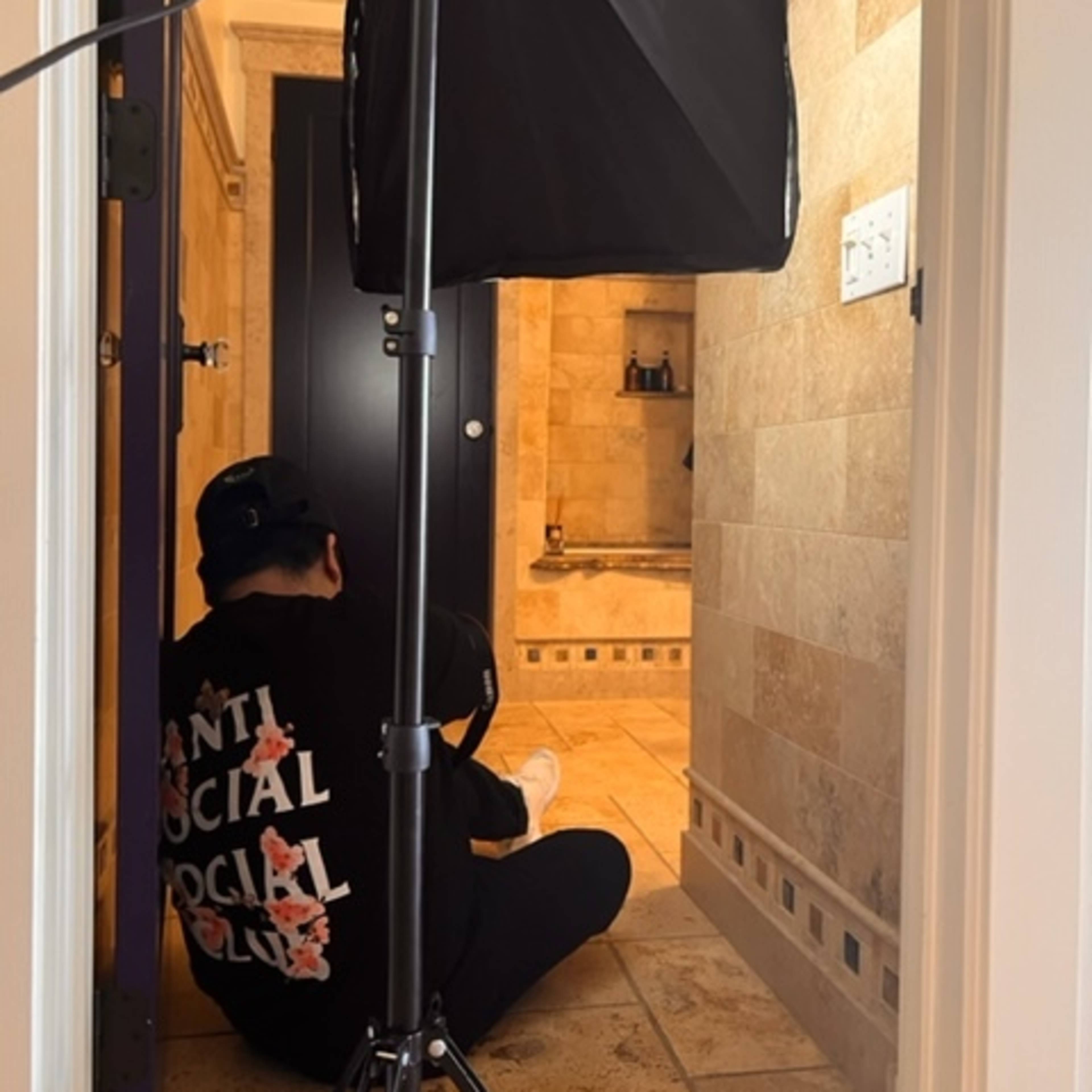 A person wearing a black sweatshirt sits on the floor in a hallway next to a lighting setup, facing an open door leading to a tiled bathroom.