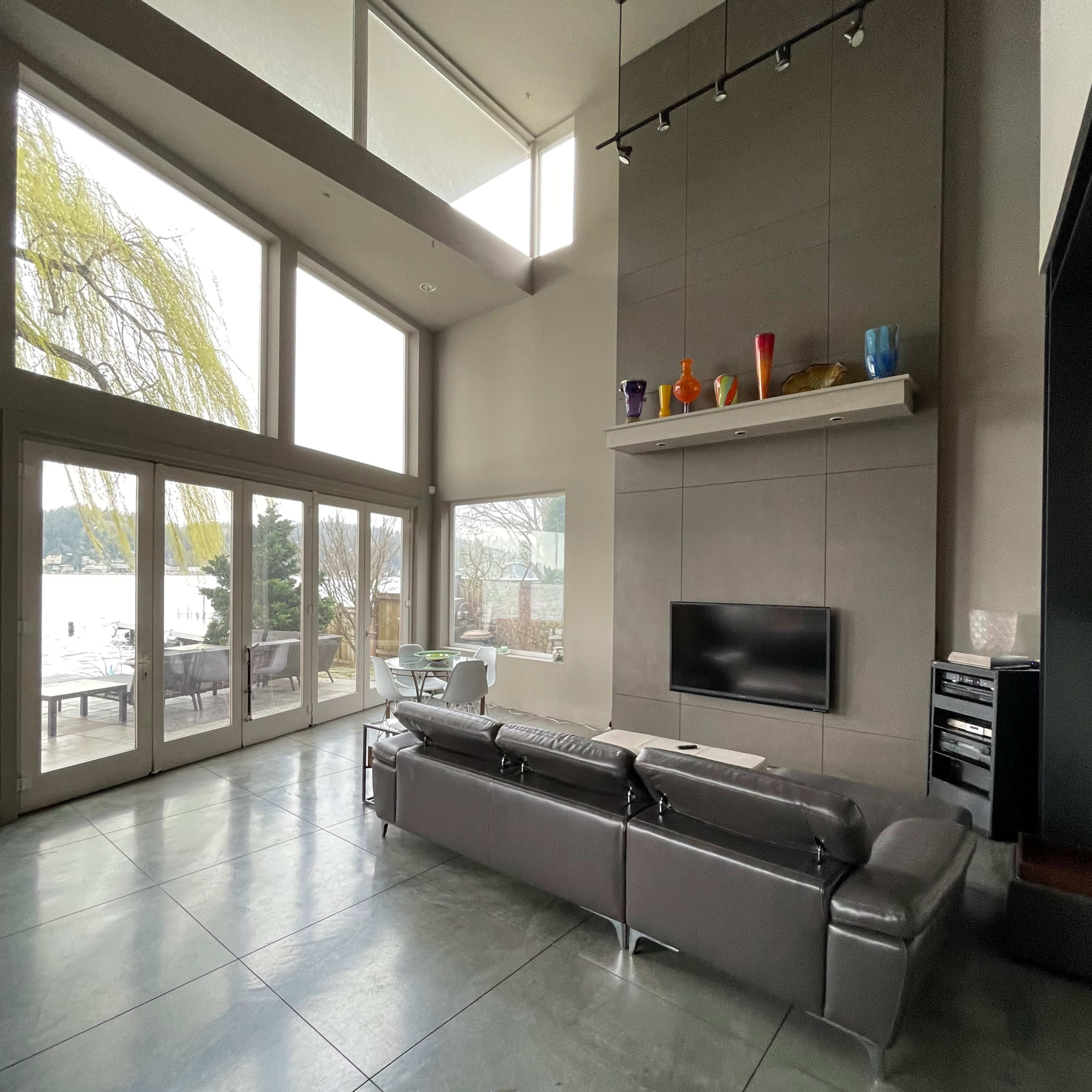 The spacious living room features large windows overlooking a lake, a modern silver couch, and a simple fireplace with colorful decor above it.