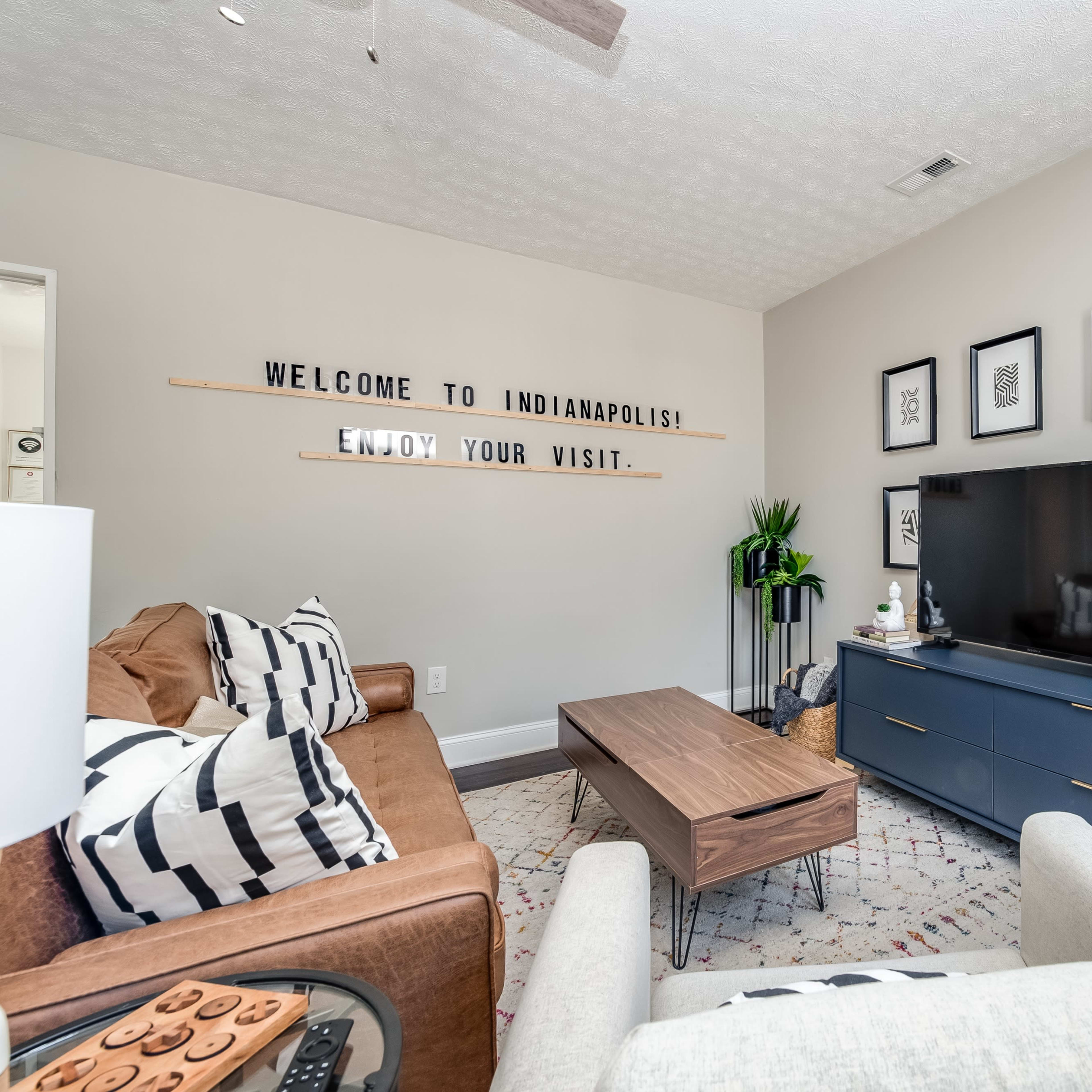 A cozy living room features a brown sofa, a gray TV stand, and framed artwork on the walls, with a sign that reads "WELCOME TO INDIANAPOLIS! ENJOY YOUR VISIT."