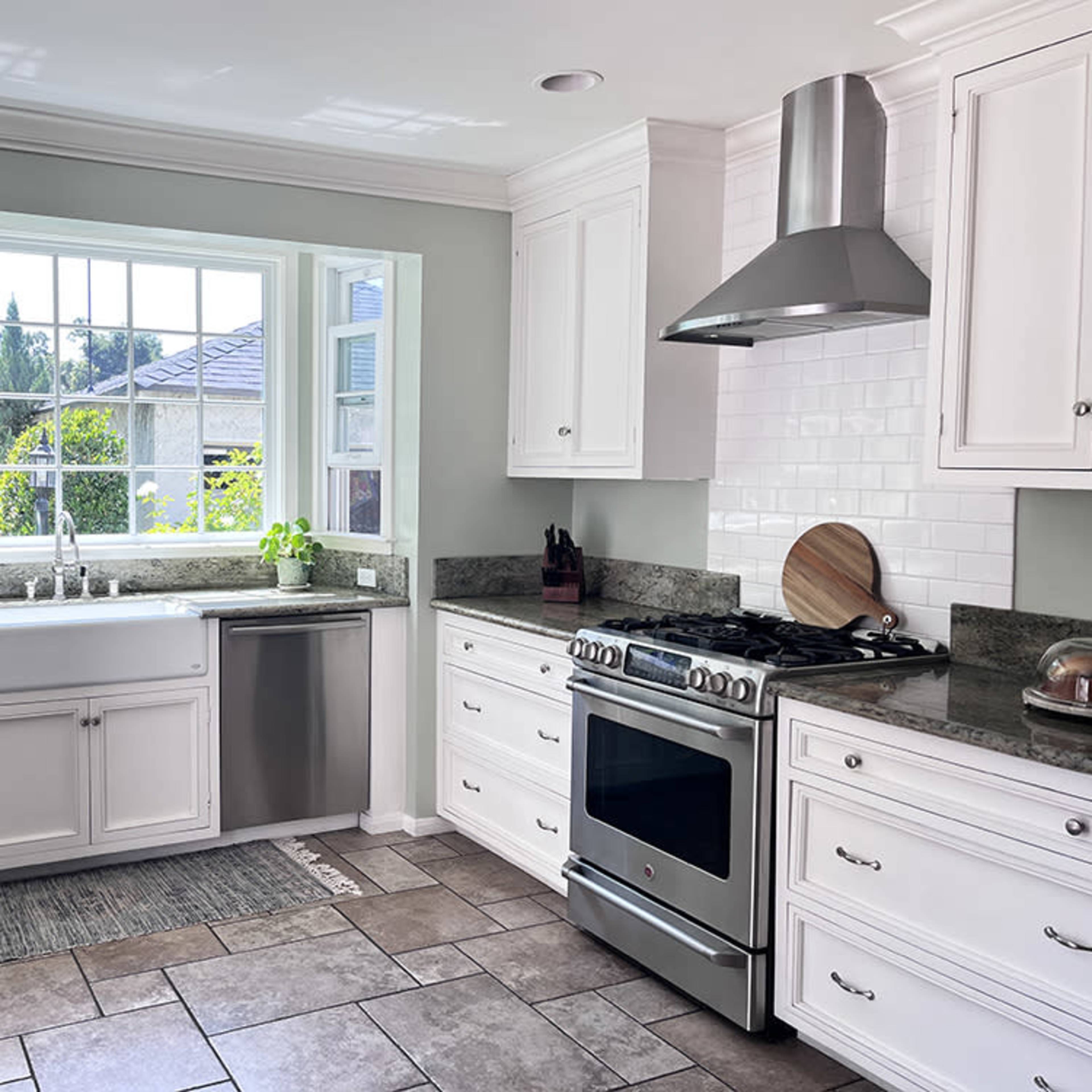 A modern kitchen features white cabinetry, a stainless steel stove, and a large window above a farmhouse sink.