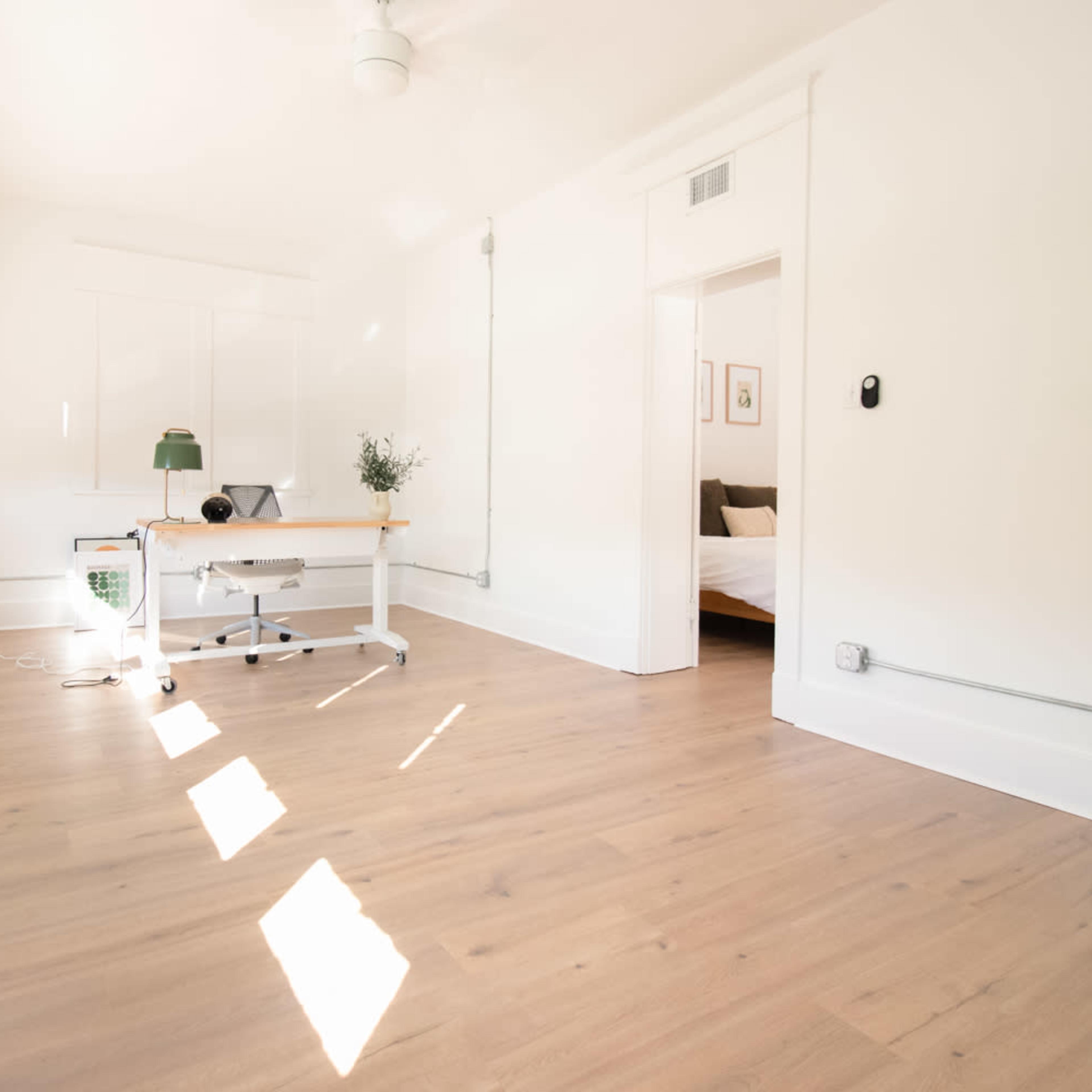 A bright, minimalist room features a wooden desk and a lamp, with sunlight streaming through several windows.