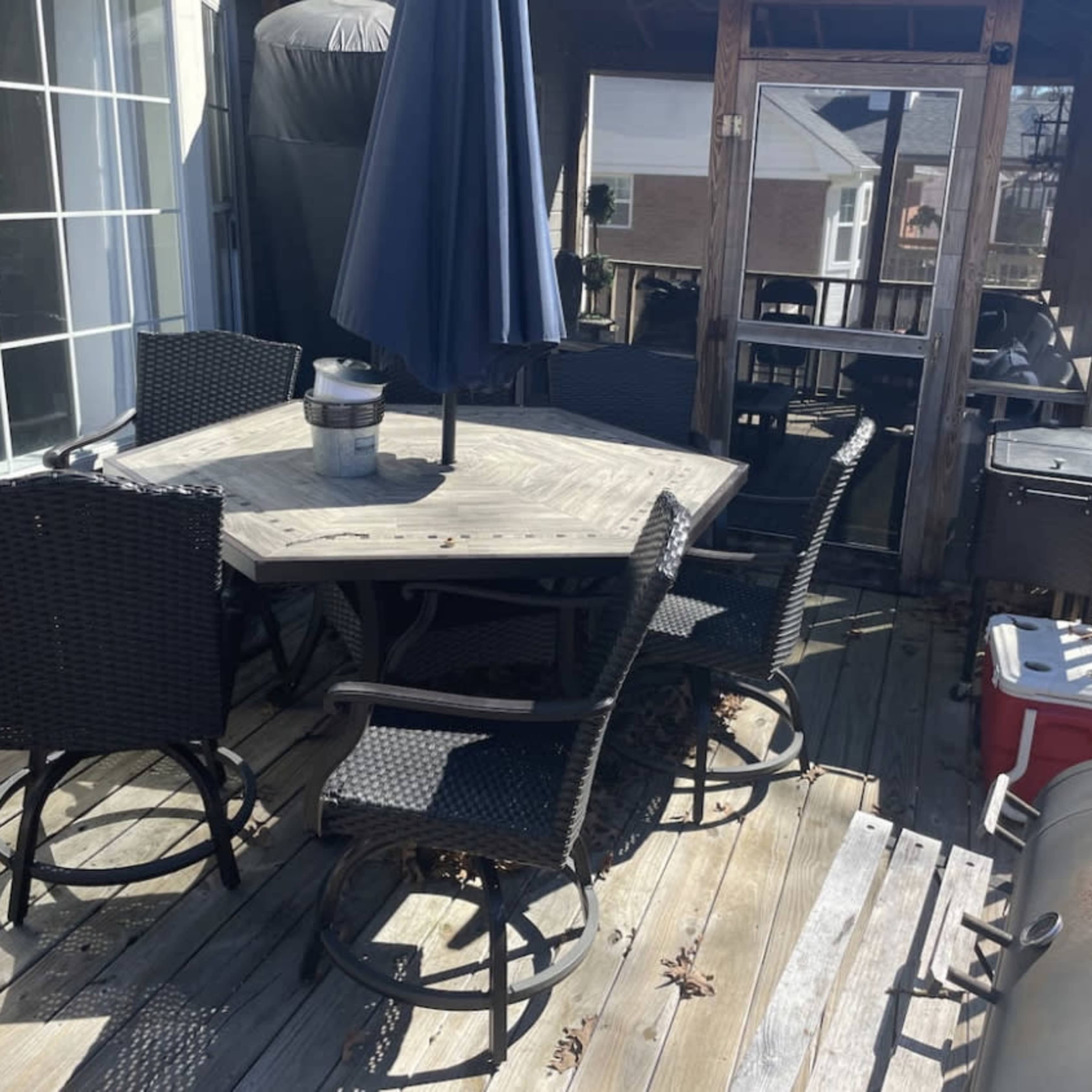 A wooden deck features an octagonal table surrounded by six wicker chairs, accompanied by a grill and a cooler in the corner.