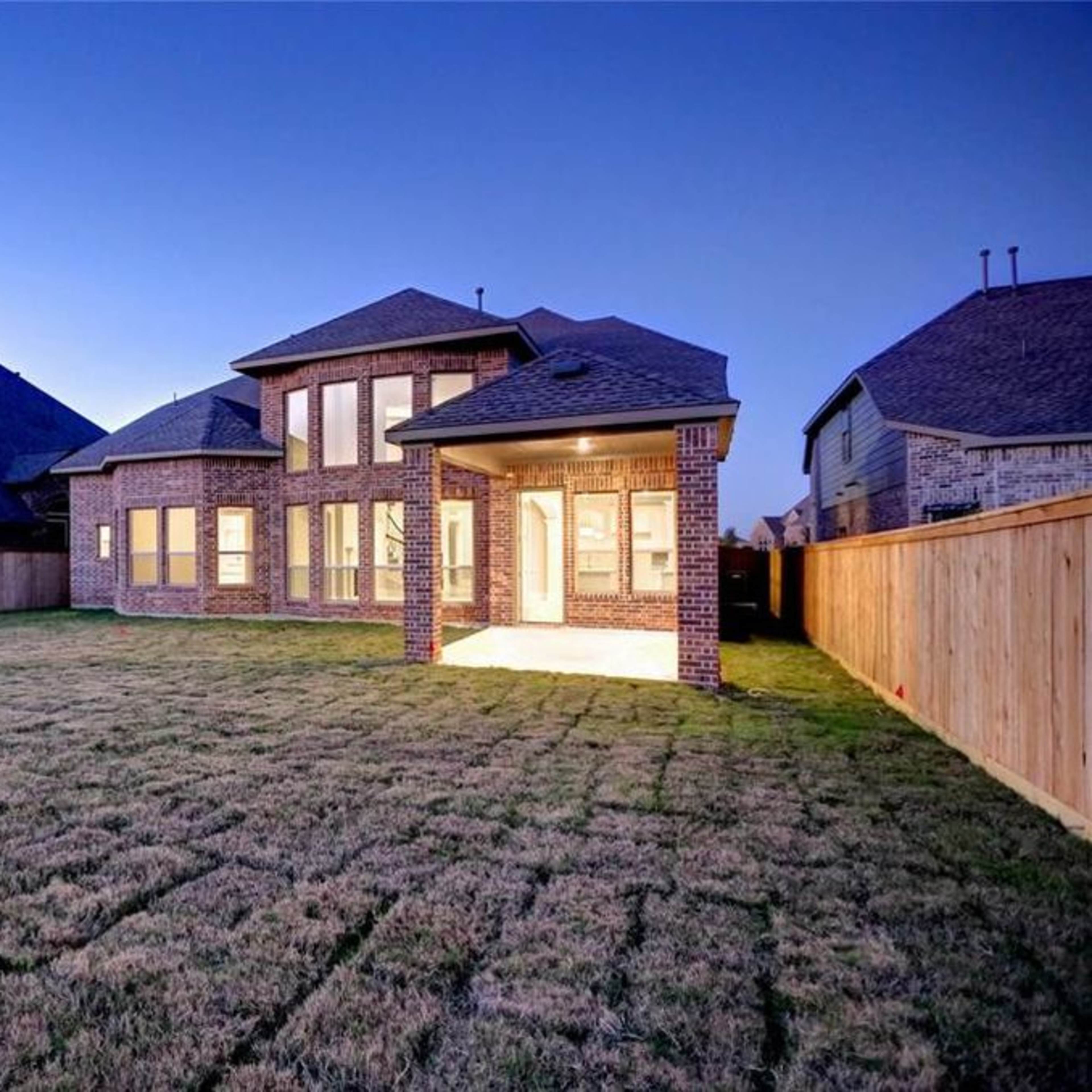 A two-story brick house with large windows is situated in a fenced backyard, illuminated at dusk.