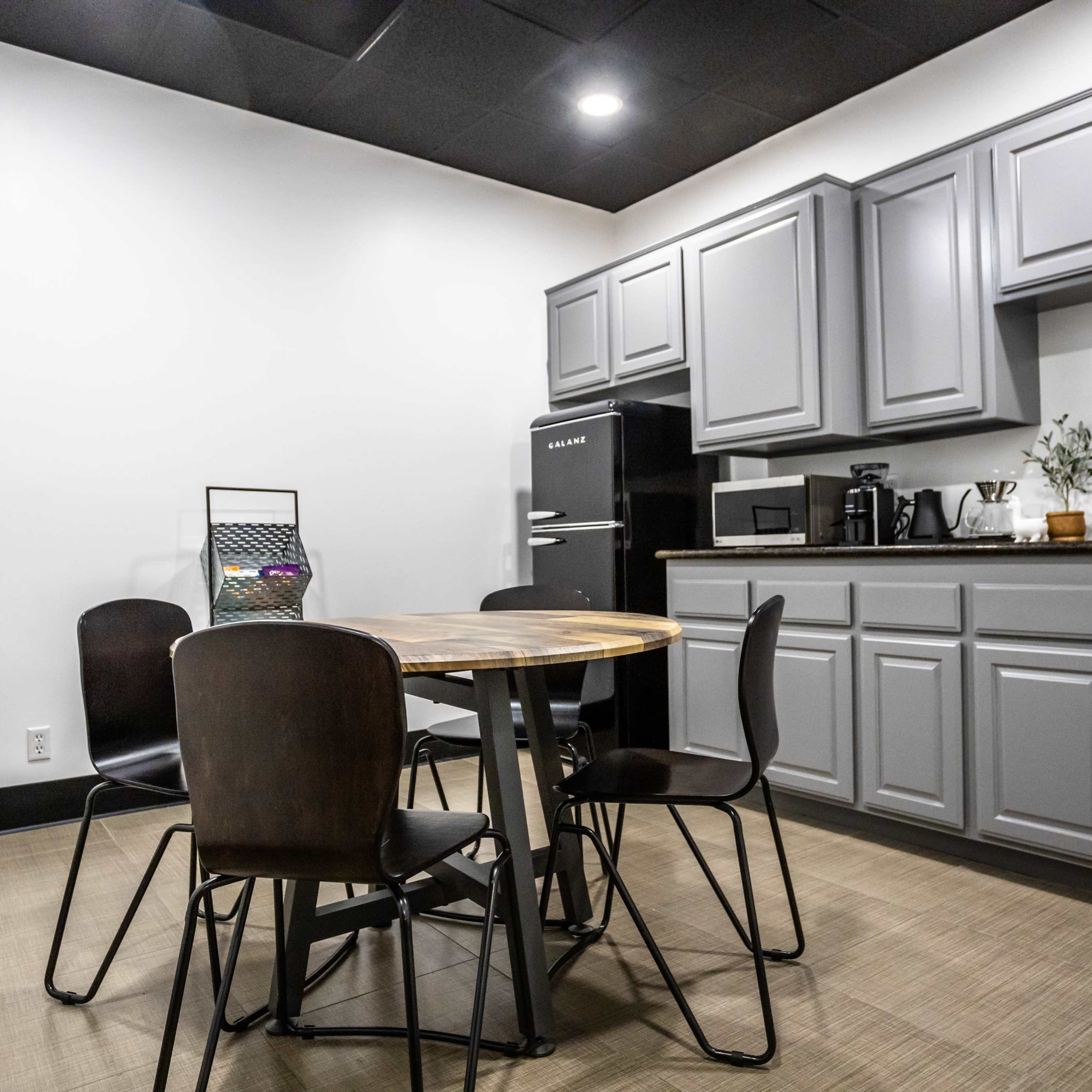 A modern kitchen features gray cabinets, a round wooden table with four chairs, and appliances including a refrigerator and coffee maker.