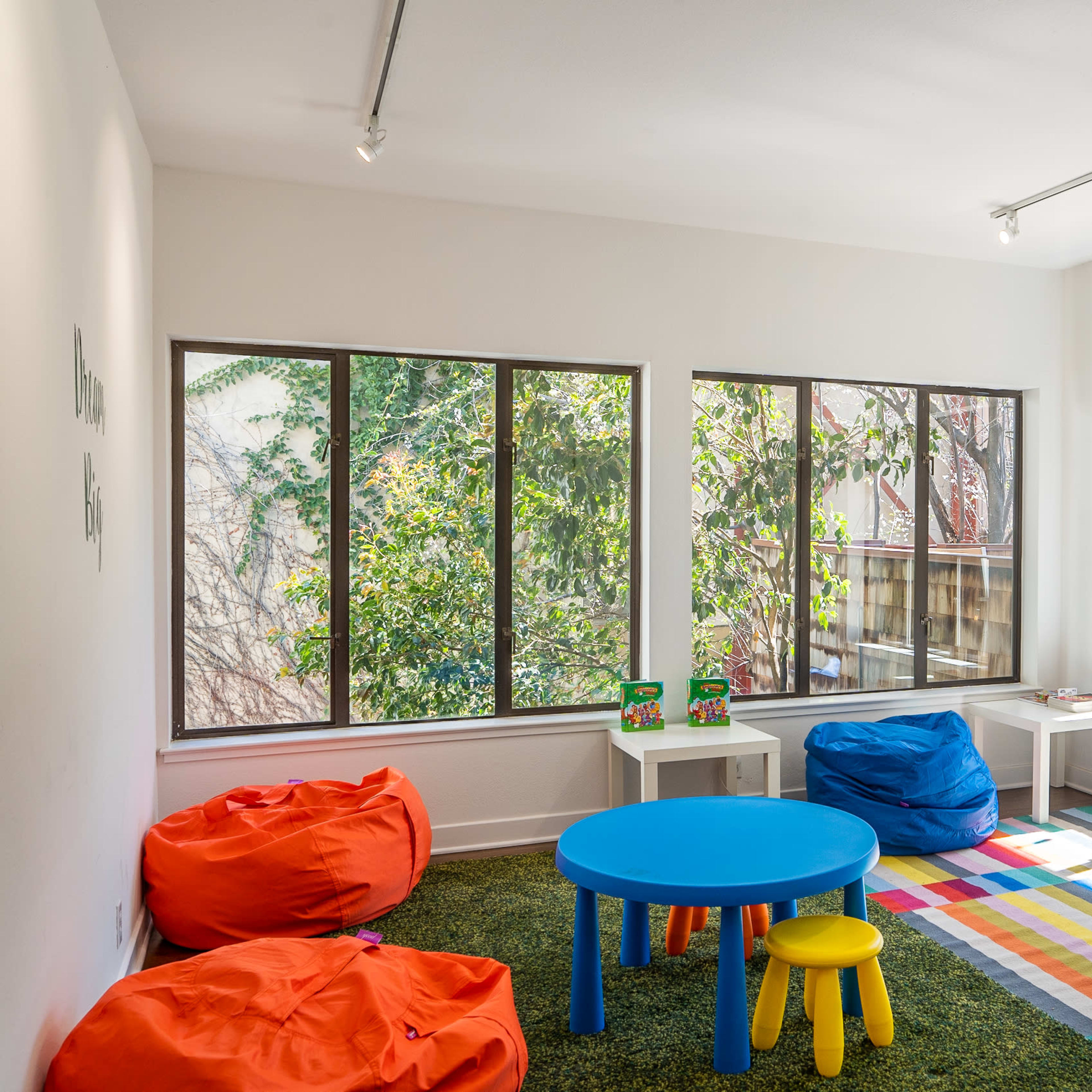 A playroom features colorful bean bags, a round blue table, and large windows that provide natural light and a view of greenery outside.