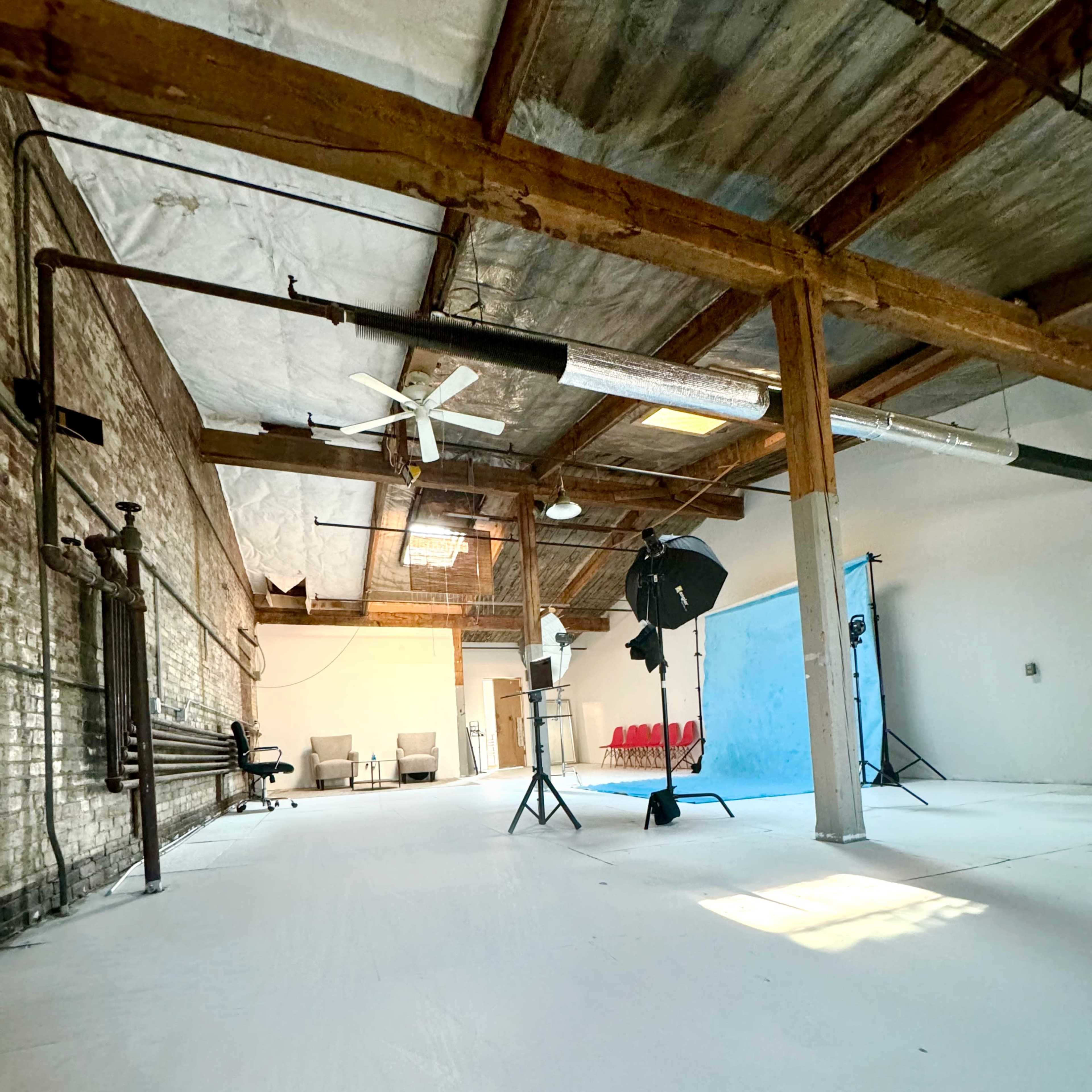 An empty studio space with white floors, exposed wooden beams, a blue backdrop, and lighting equipment arranged in the corner.