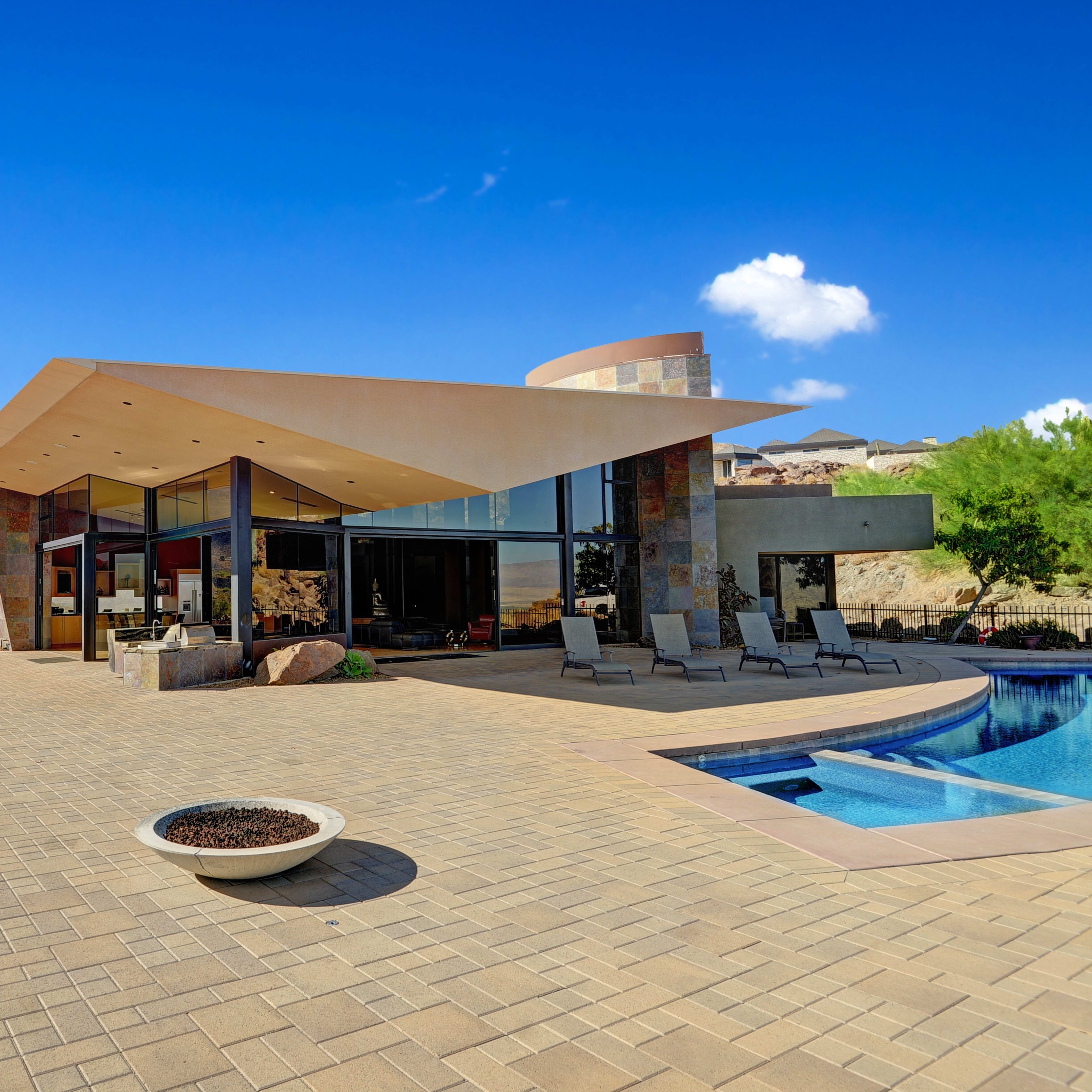 A modern, angular house is situated near a swimming pool, surrounded by rocky terrain and mountains under a clear blue sky.
