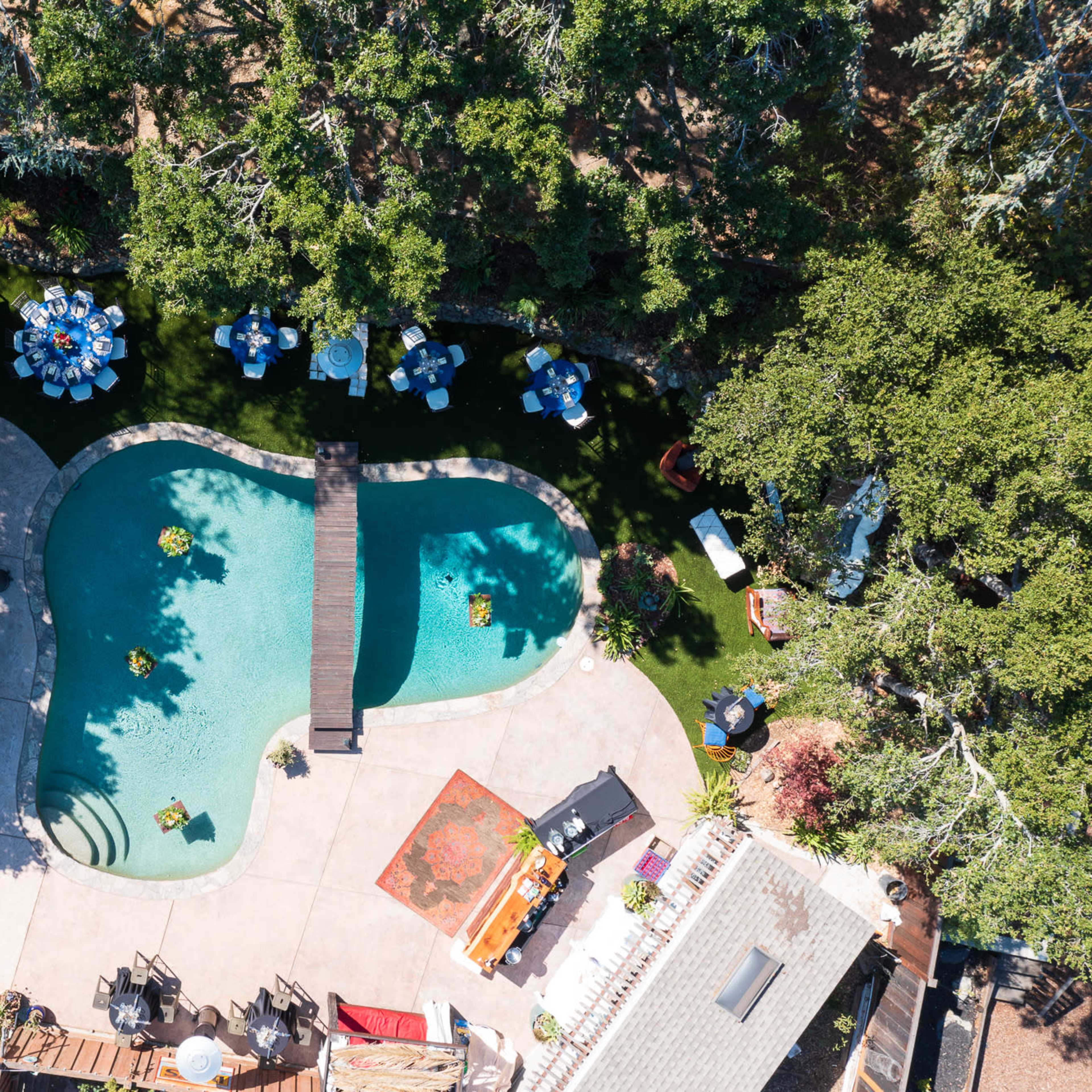An aerial view of a landscaped backyard featuring a curvy swimming pool, a wooden bridge, and several tables arranged around it.