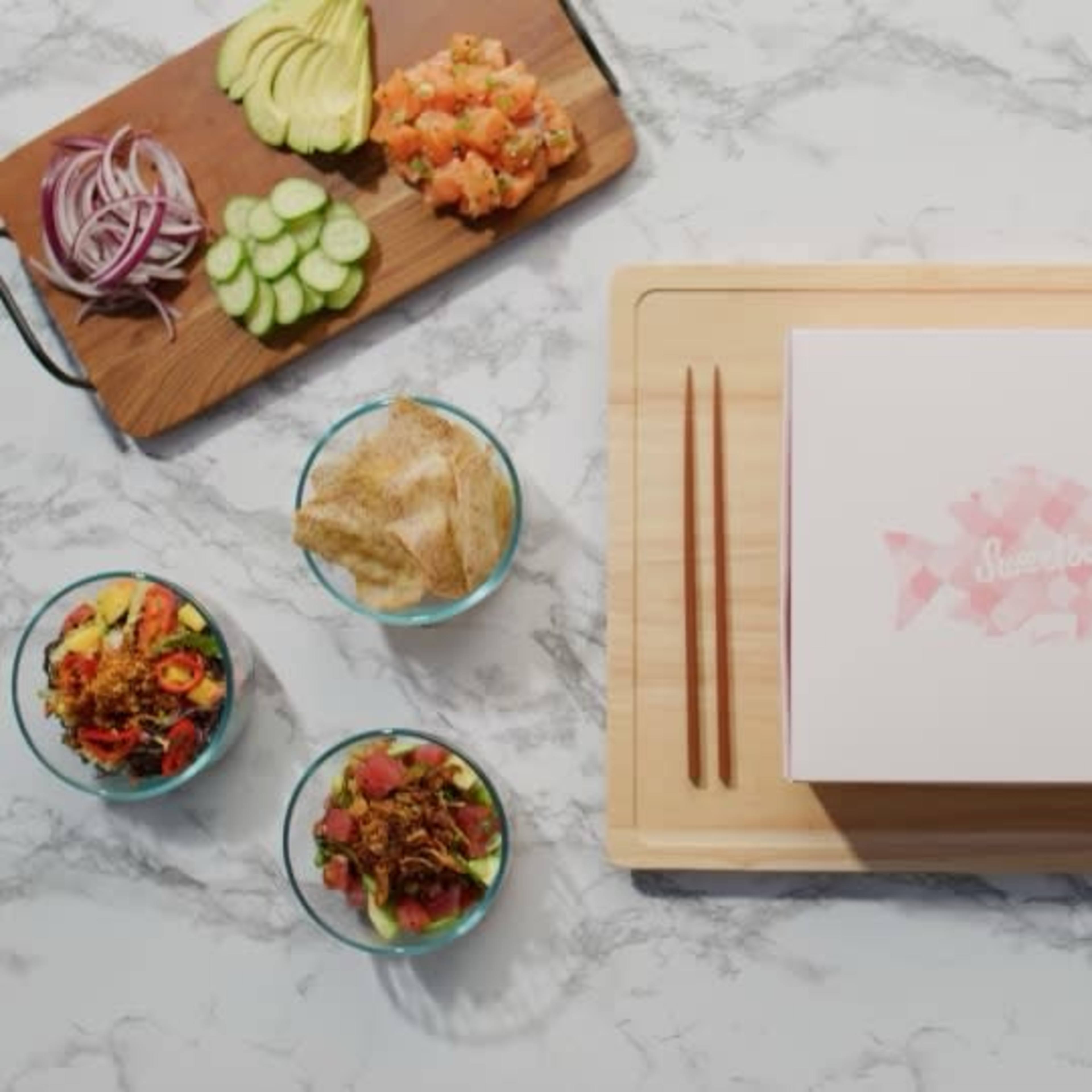 A wooden cutting board with a pink fish-patterned box at the center, surrounded by various dishes of fresh ingredients and toppings, including sliced vegetables and seasonings.