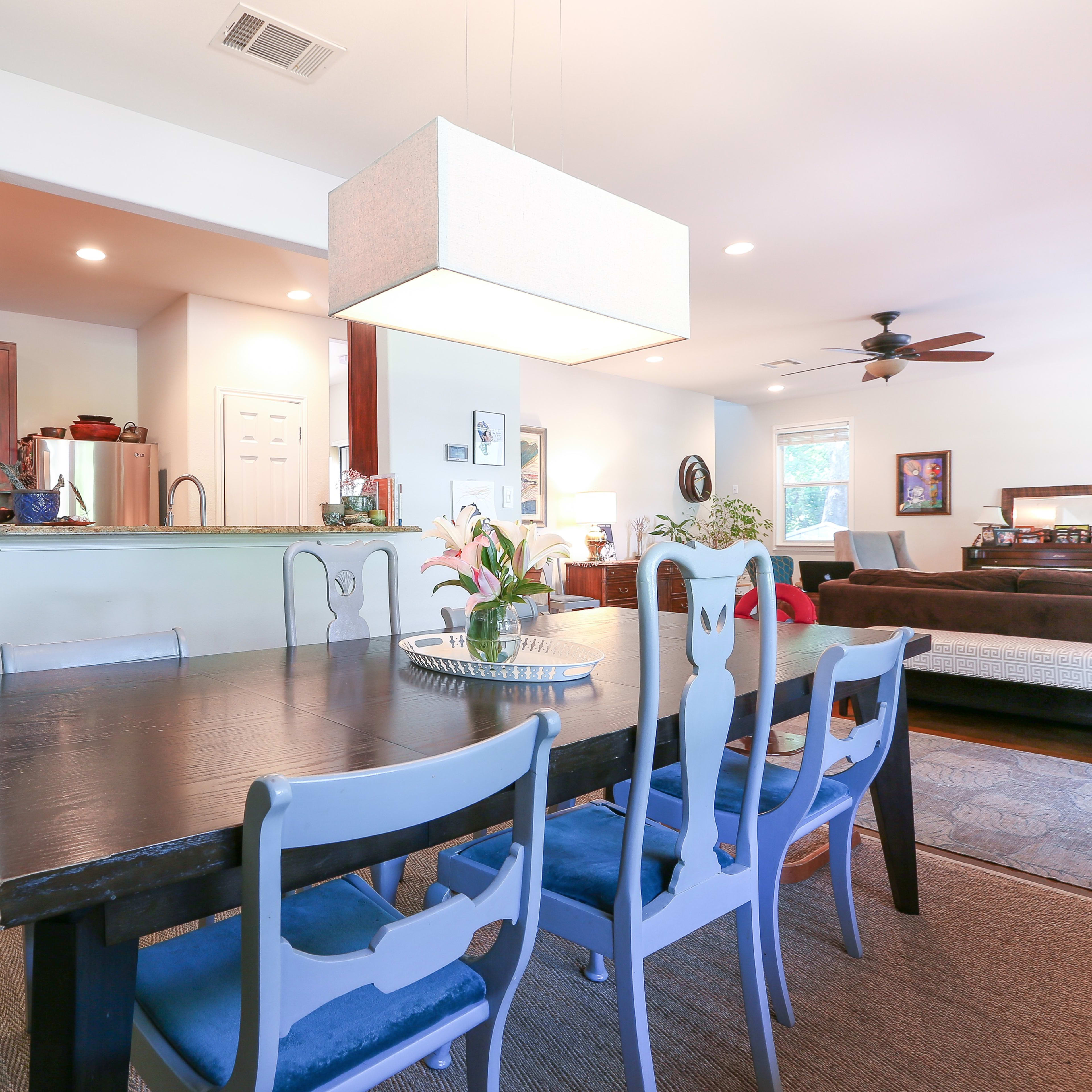 A dining area with a long wooden table surrounded by blue upholstered chairs, and a light fixture hanging above it, with an open kitchen and living room visible in the background.