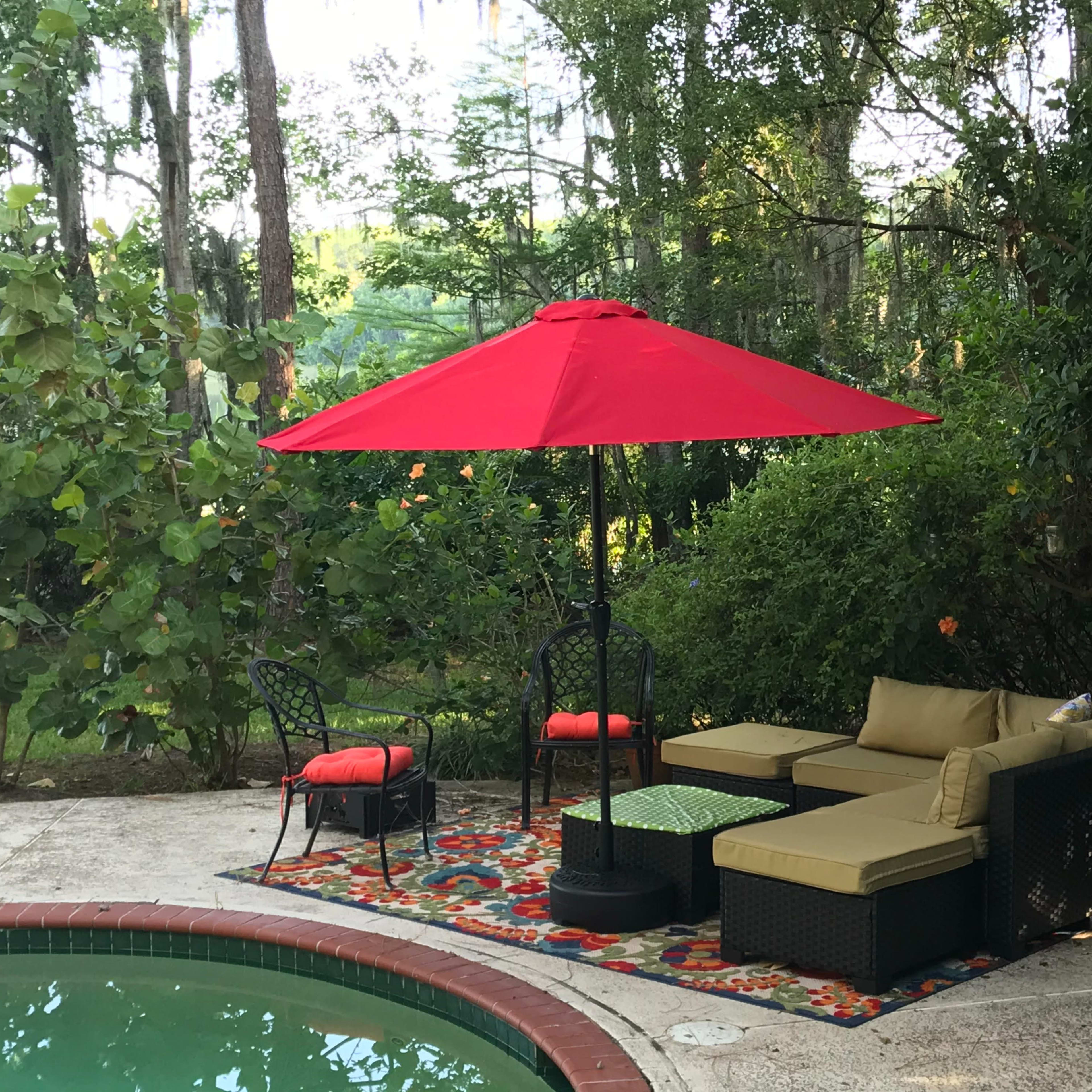 A red umbrella shades a seating area with a sofa and chairs beside a pool surrounded by greenery.