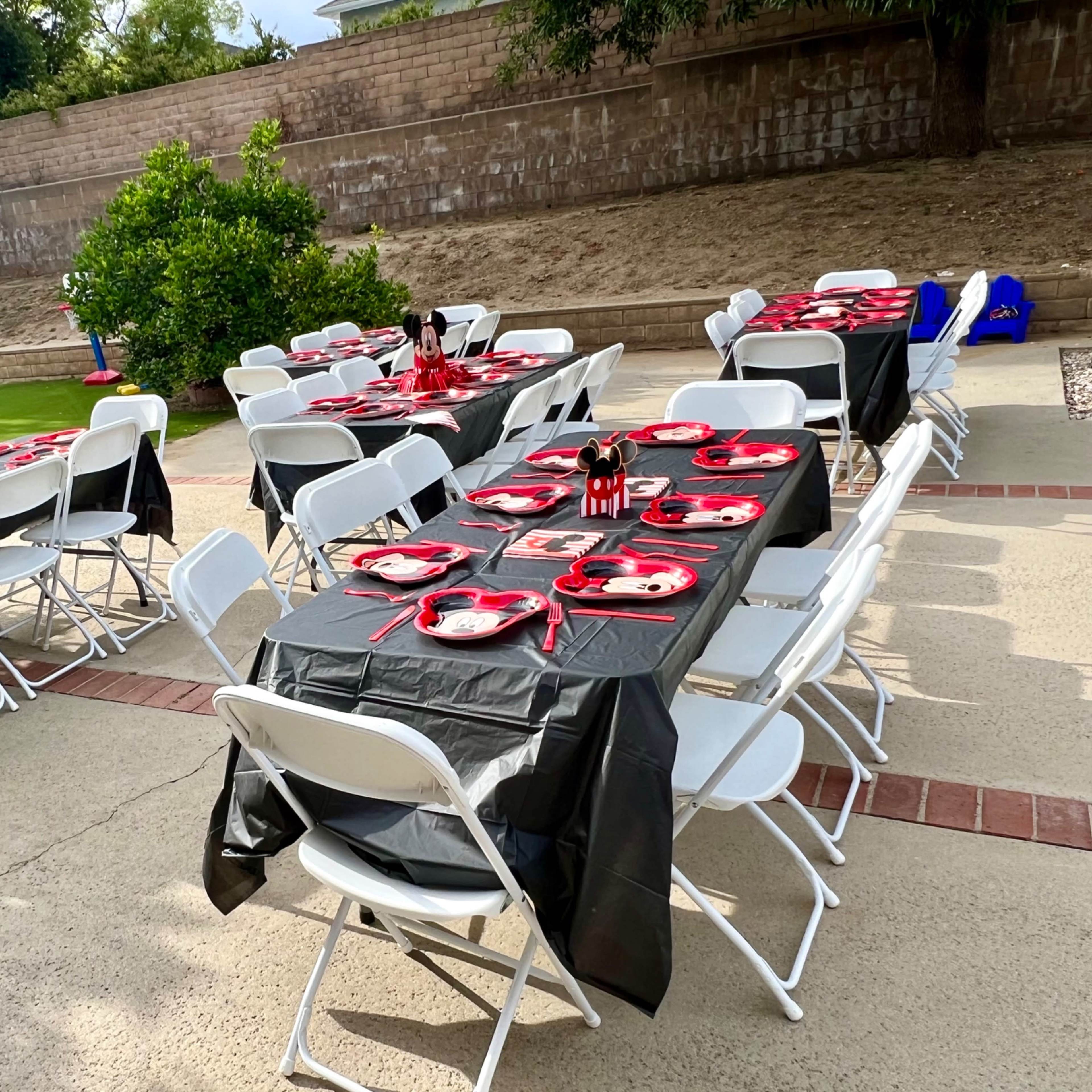 The scene features several tables set up outdoors with black tablecloths and red plates, arranged for a dining event.