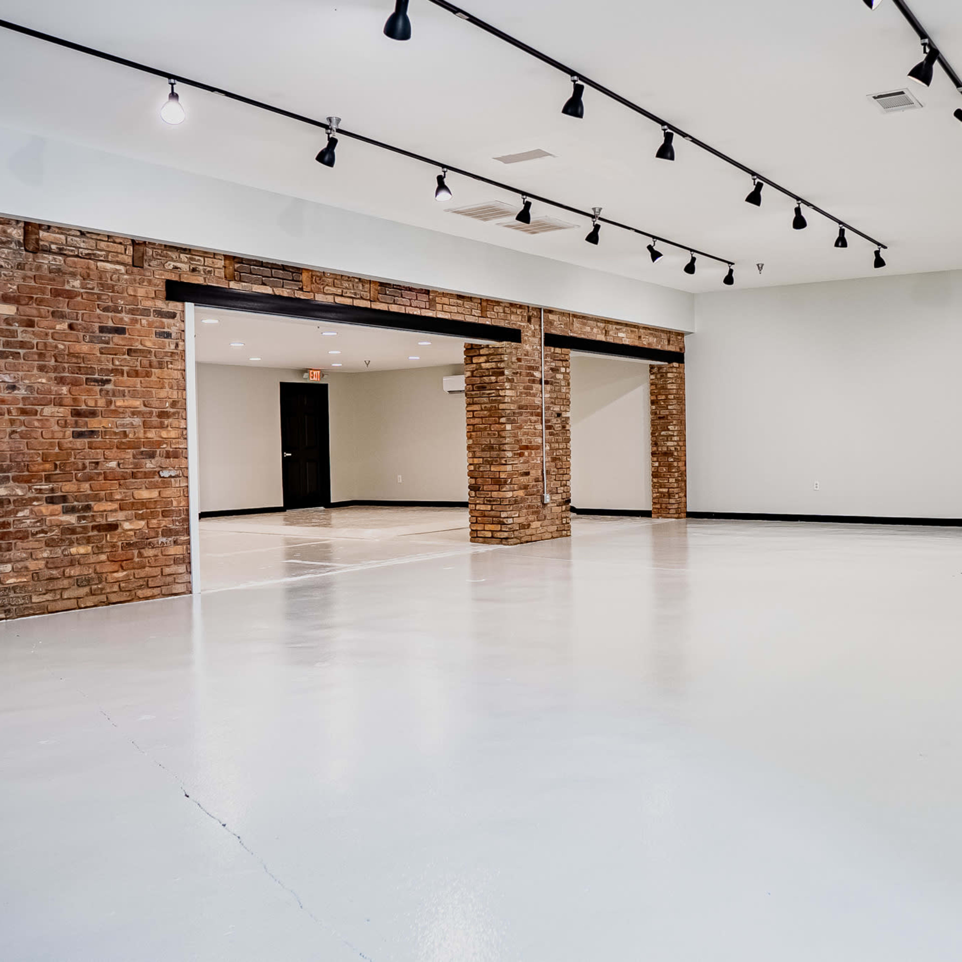 A spacious, empty room with white floors, exposed brick columns, and track lighting on the ceiling.