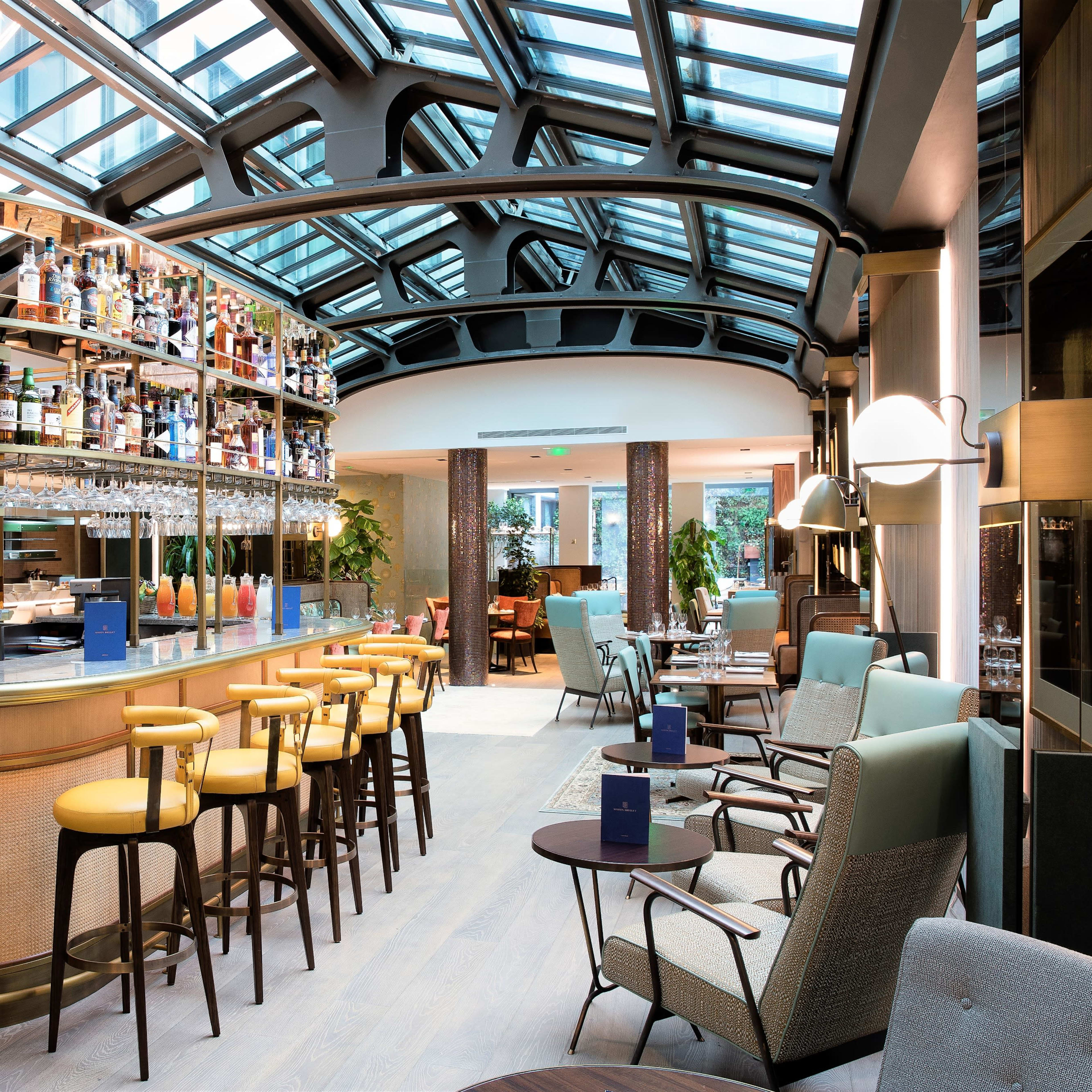 A modern bar area inside a well-lit restaurant, featuring a glass ceiling and shelves stocked with various bottles.