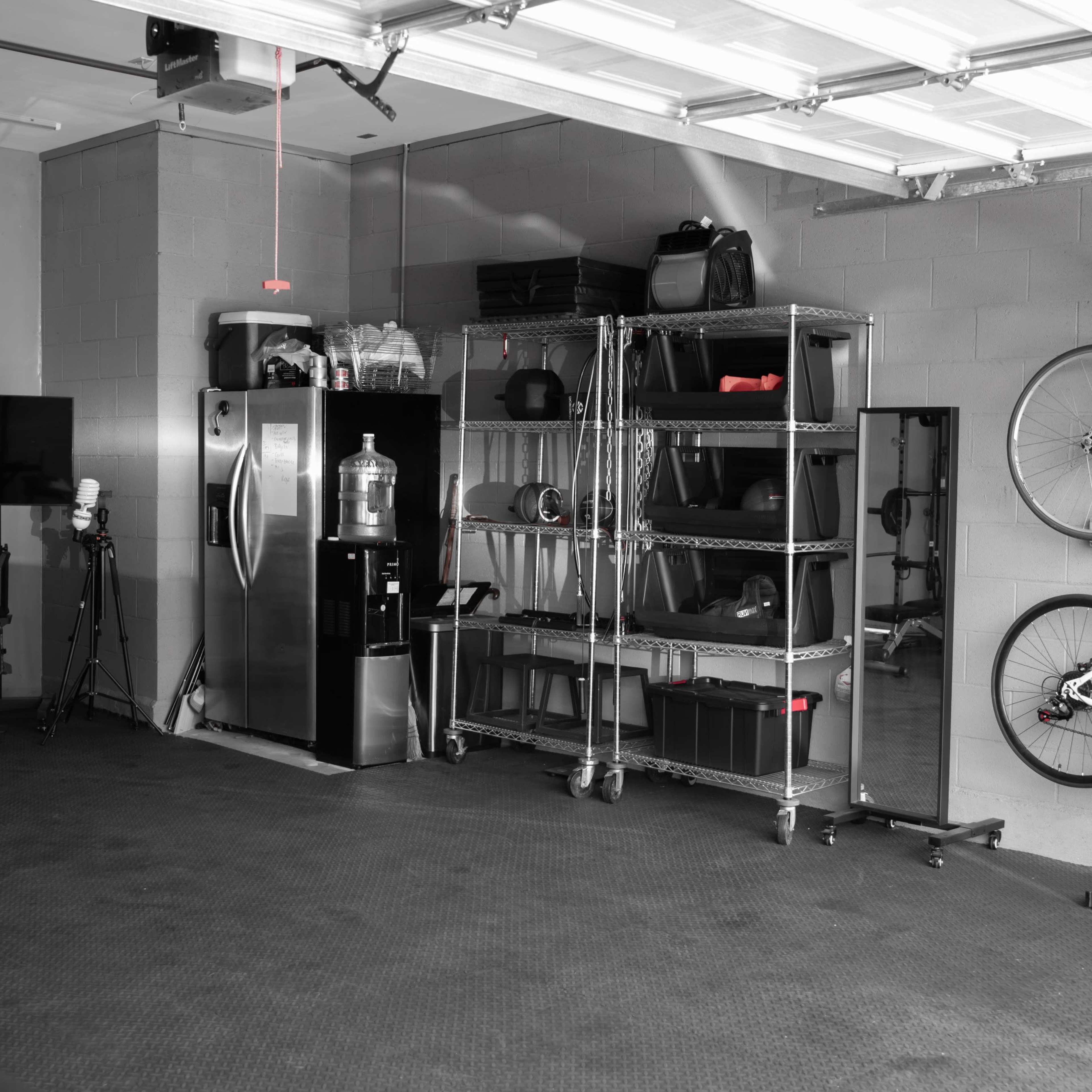 A spacious garage features a refrigerator, a water cooler, a shelving unit with various items, a stationary bike, and two bicycles mounted on the wall.