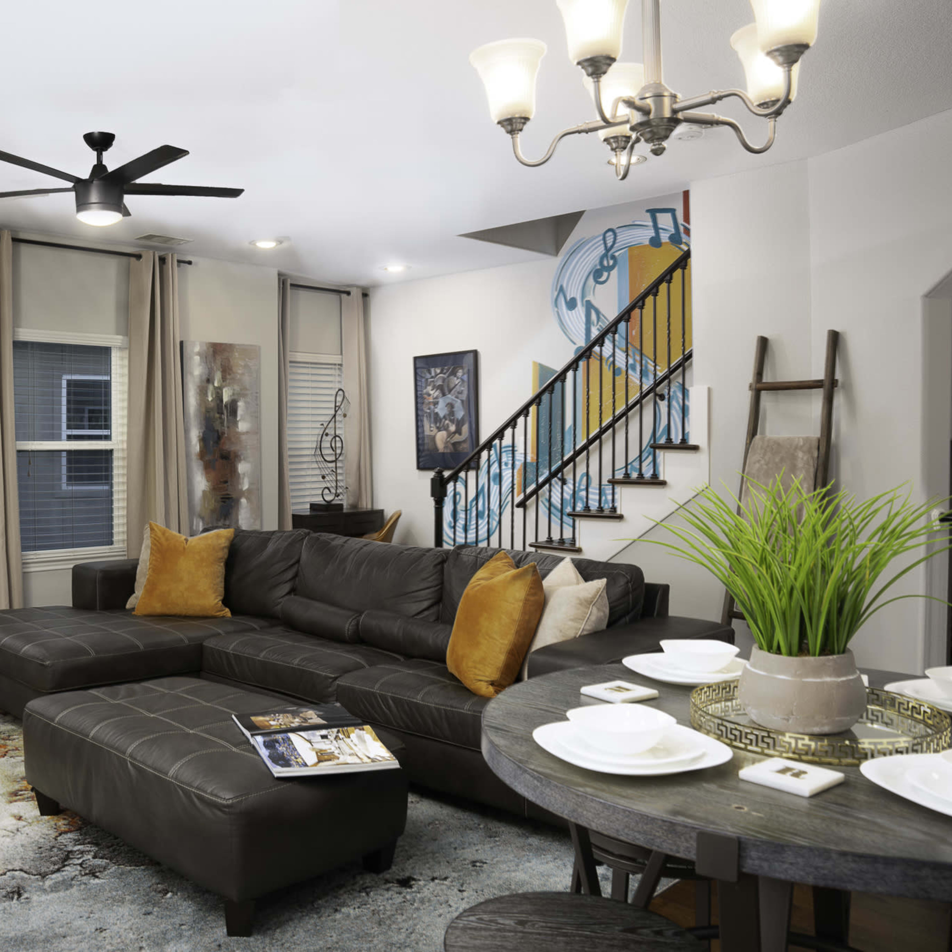A modern living room features a dark leather sectional sofa, a round dining table, and a staircase with a colorful mural.