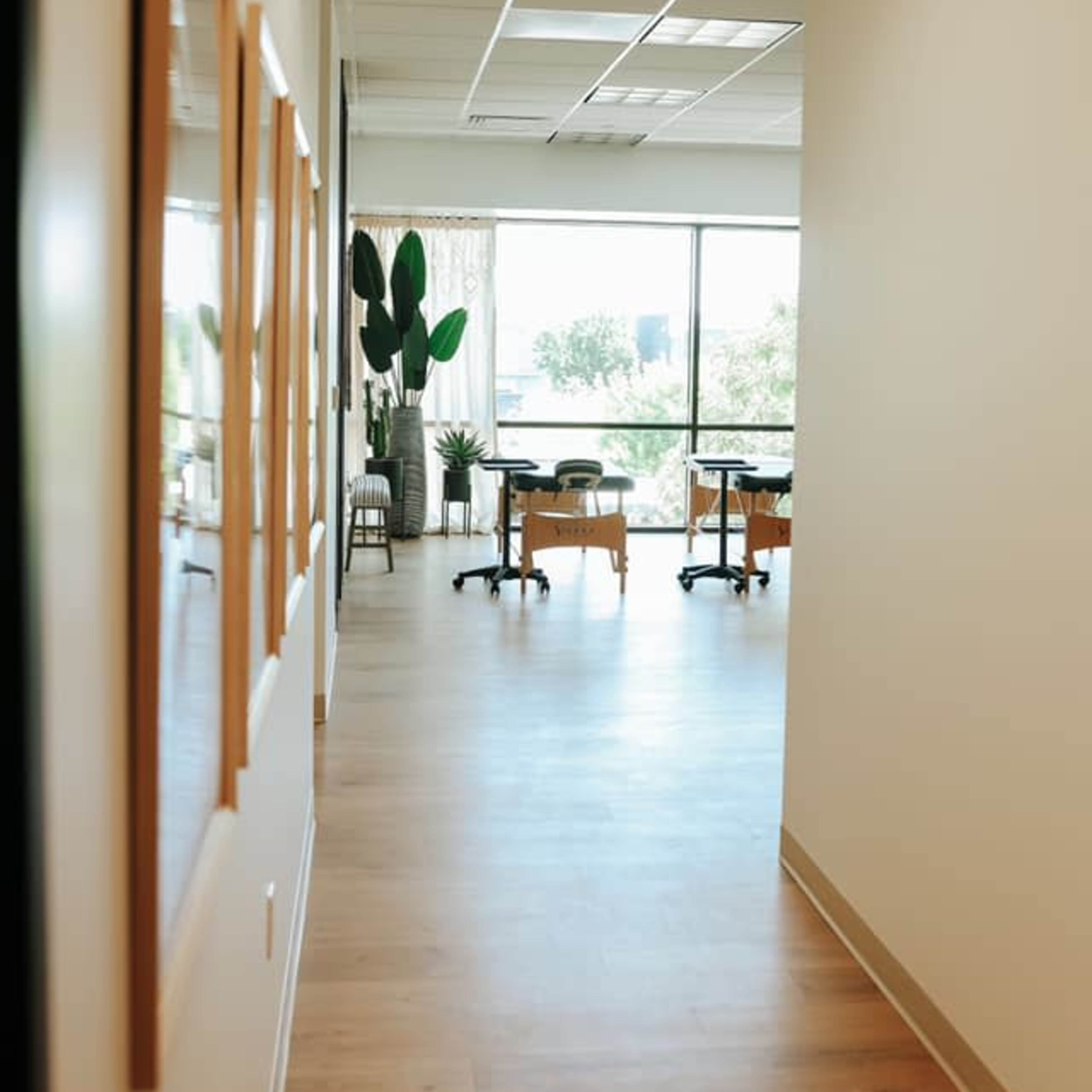 A well-lit hallway leading to a room with tables and plants, framed by large windows.