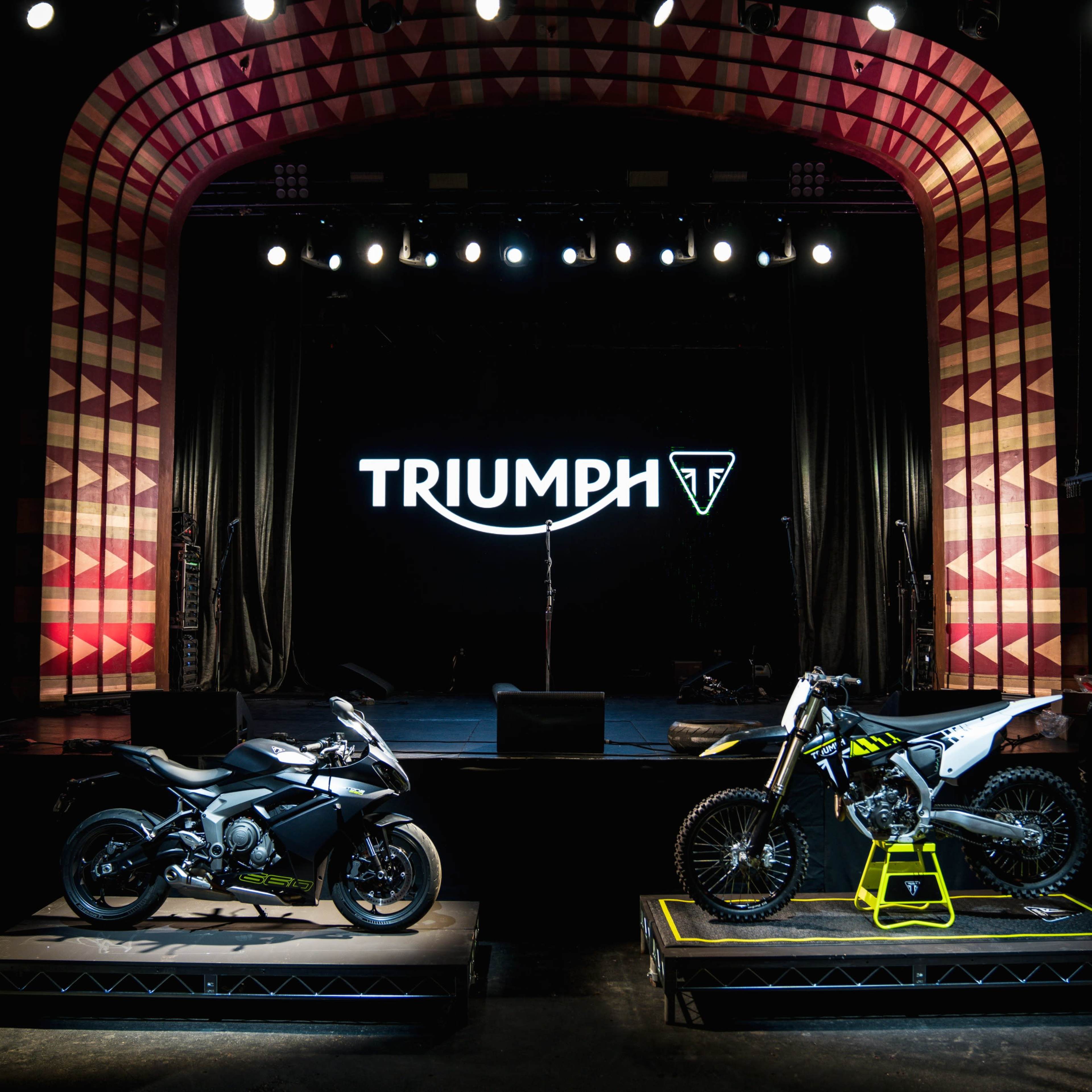 Two Triumph motorcycles displayed on platforms in a dimly lit stage setting, with a large Triumph logo illuminated in the background.