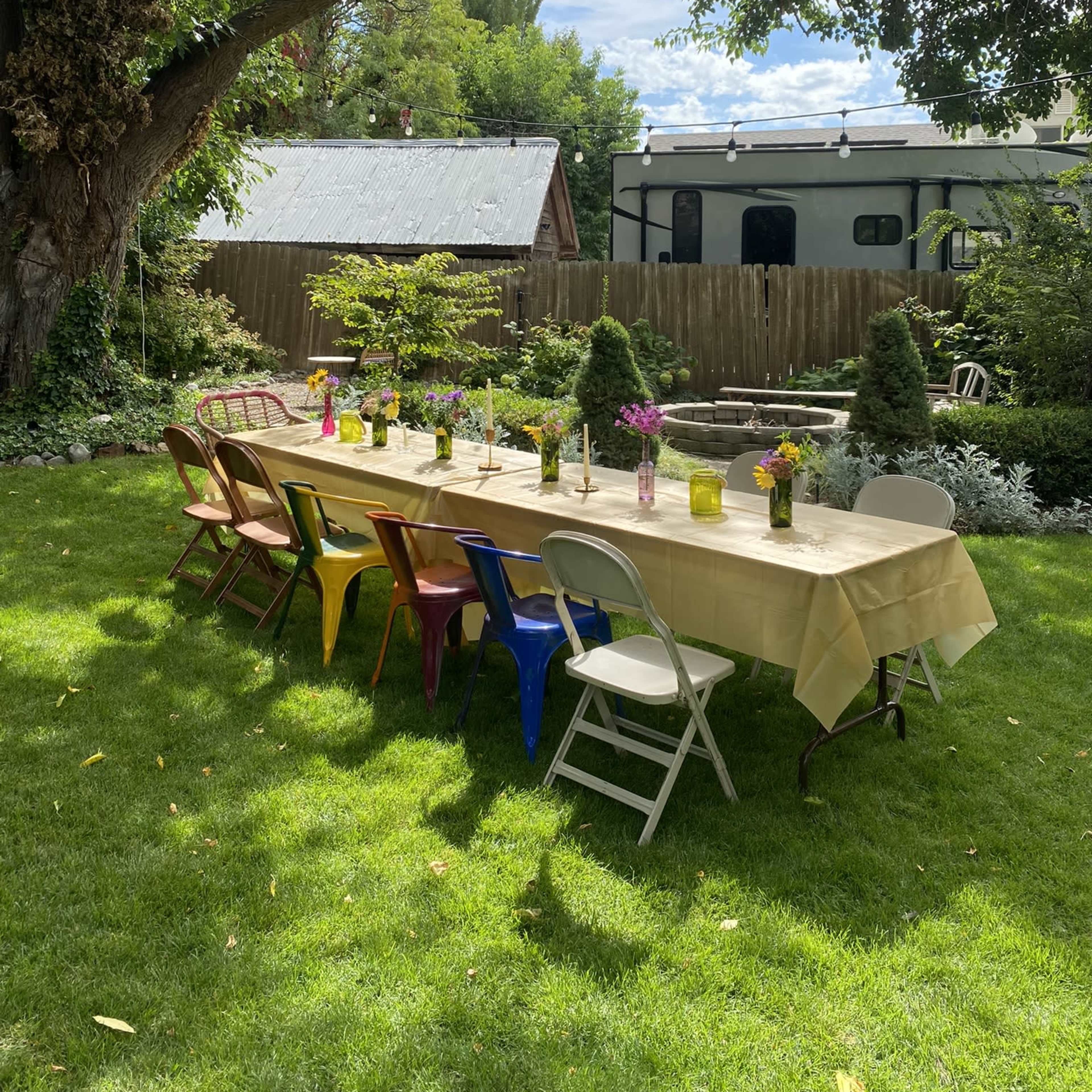 A long table is set with colorful chairs on a green lawn surrounded by trees and plants, with a backdrop of a house and trailer.