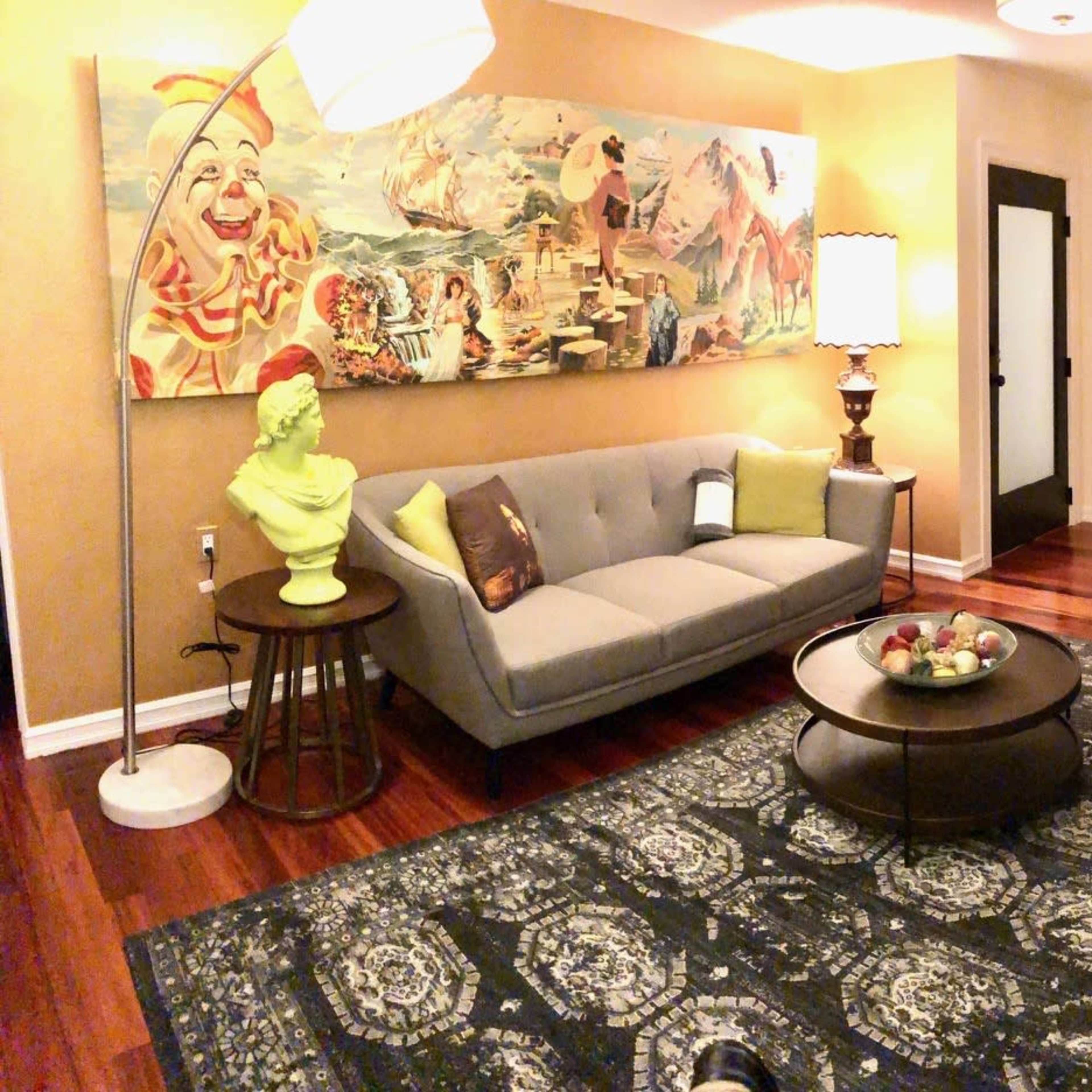 A living room featuring a gray sofa, a grand piano, colorful artwork on the wall, and a round coffee table topped with fruit.