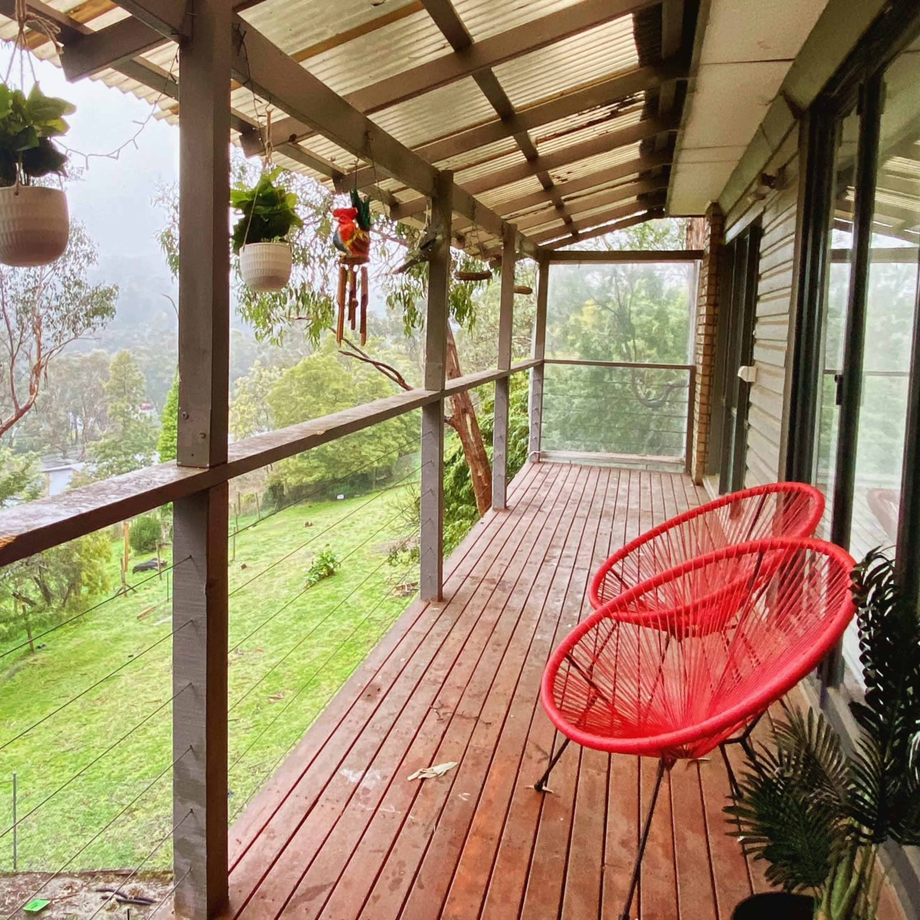 A wooden porch with two red hanging chairs, surrounded by potted plants and a view of a green landscape beyond.