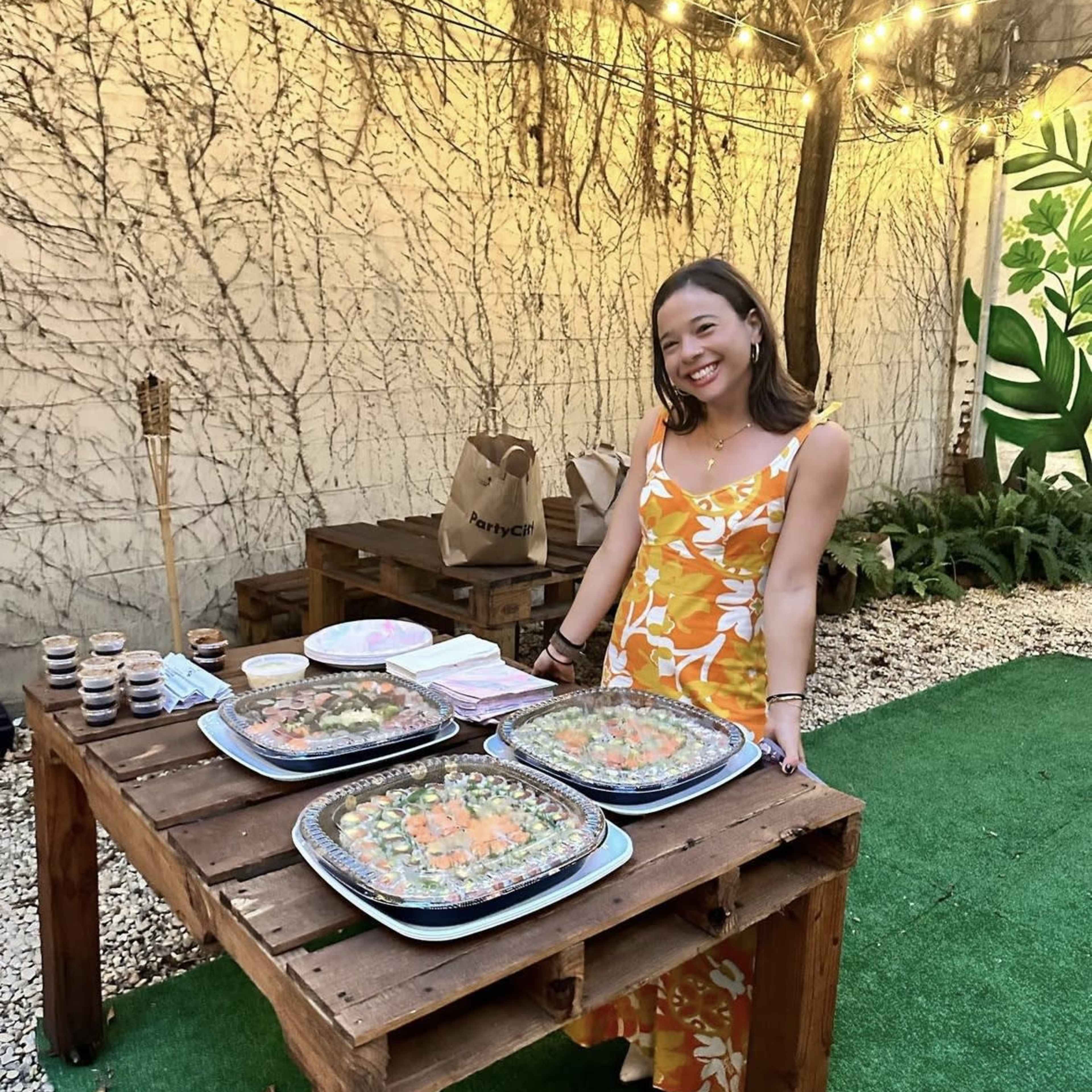 A woman stands by a wooden table outdoors, smiling as she showcases trays of sushi and small desserts in a garden setting.