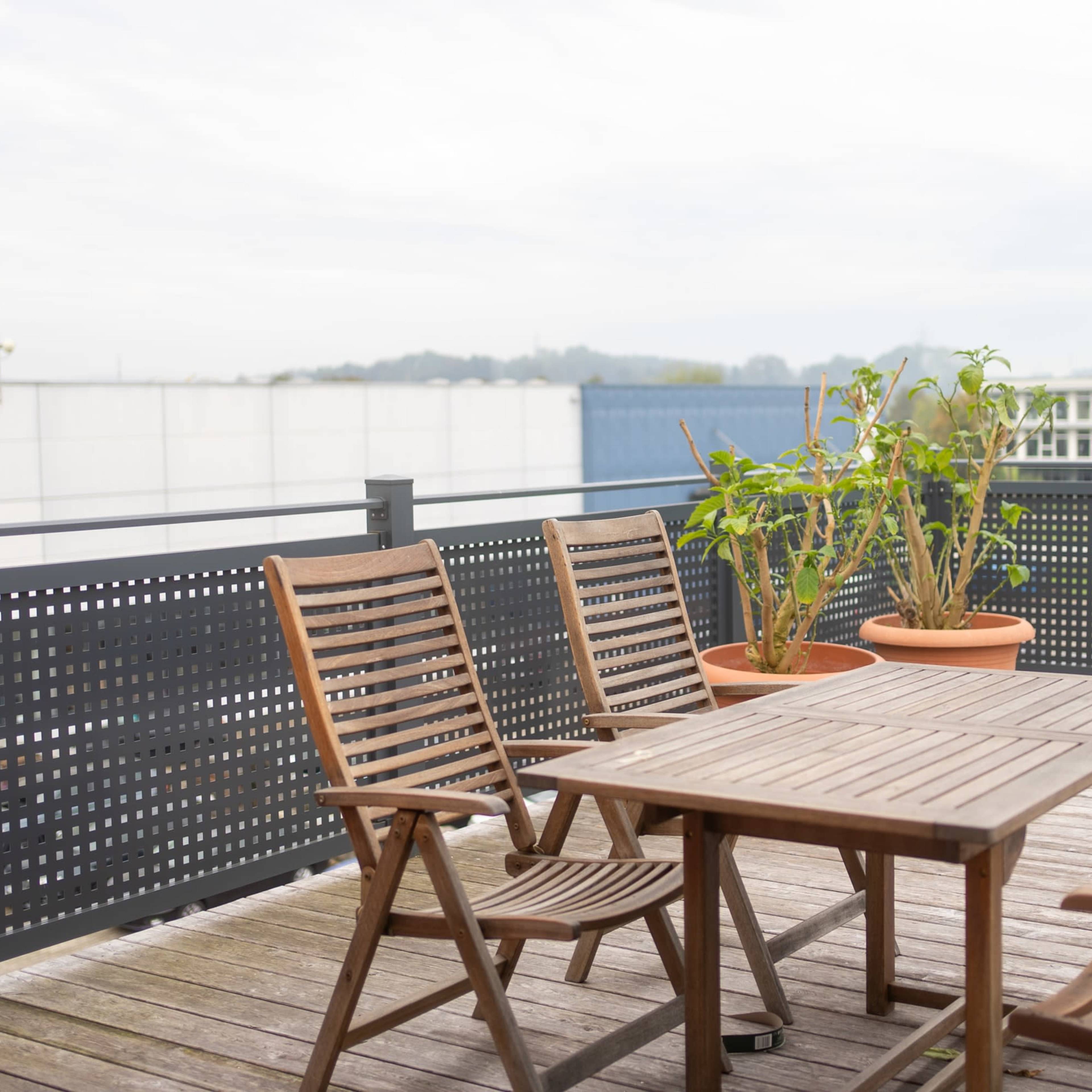A wooden patio set with a table and chairs is positioned on a deck, overlooking potted plants and a distant urban landscape.