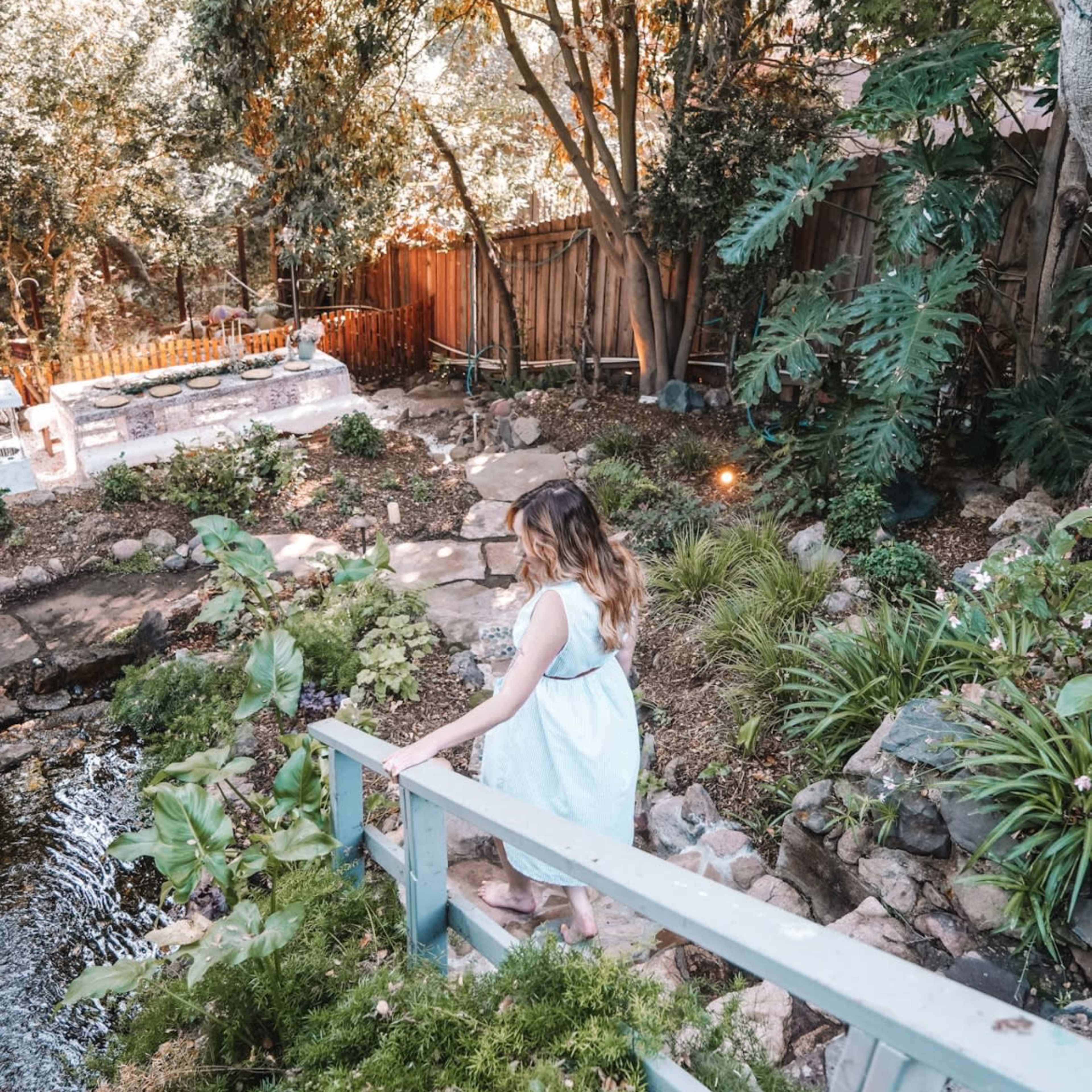 A girl walks along a wooden bridge in a landscaped garden by a pond surrounded by greenery.