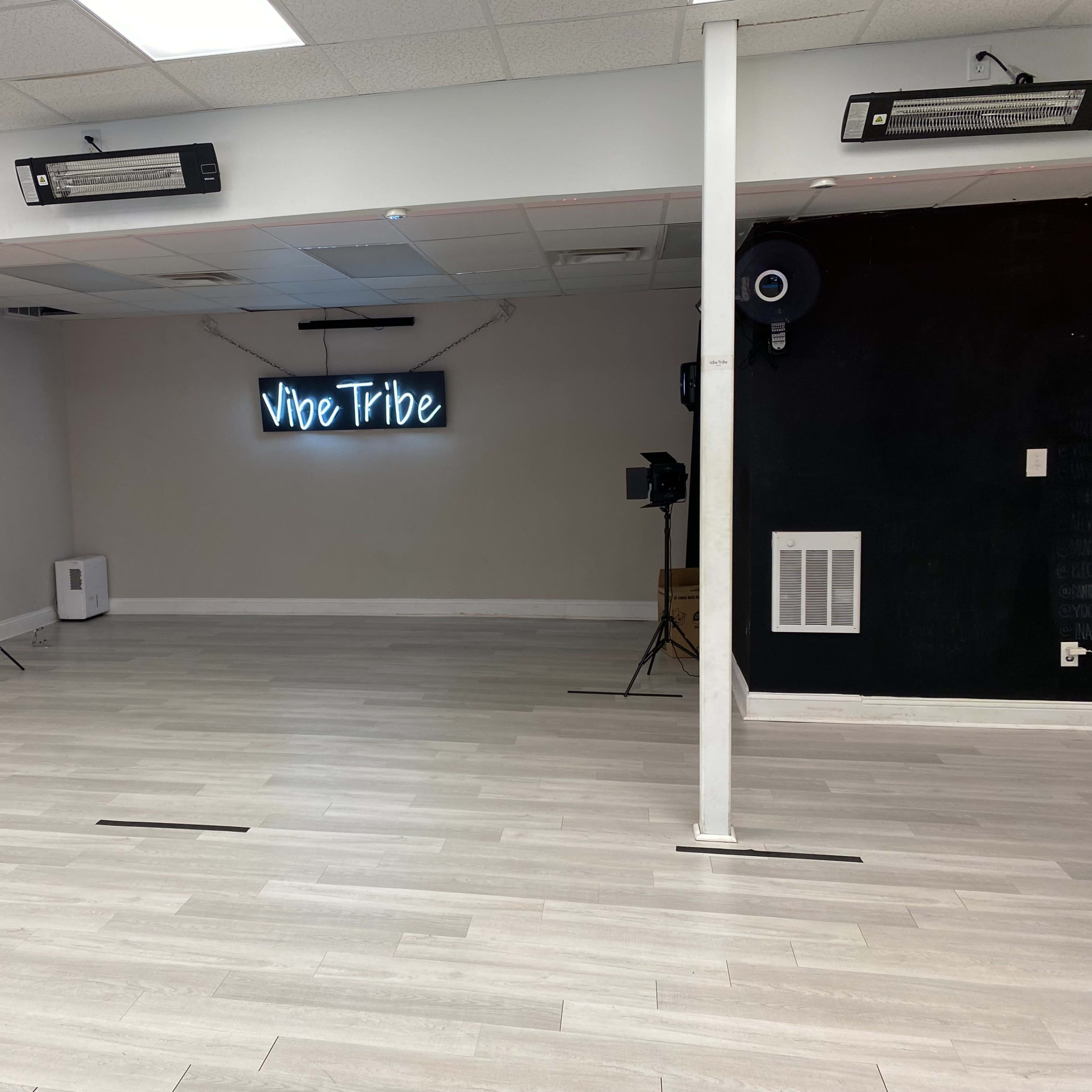 A spacious, empty room with light gray flooring and a neon sign that reads "Vibe Tribe" on the wall, along with several light fixtures and cameras set up for a possible video shoot.