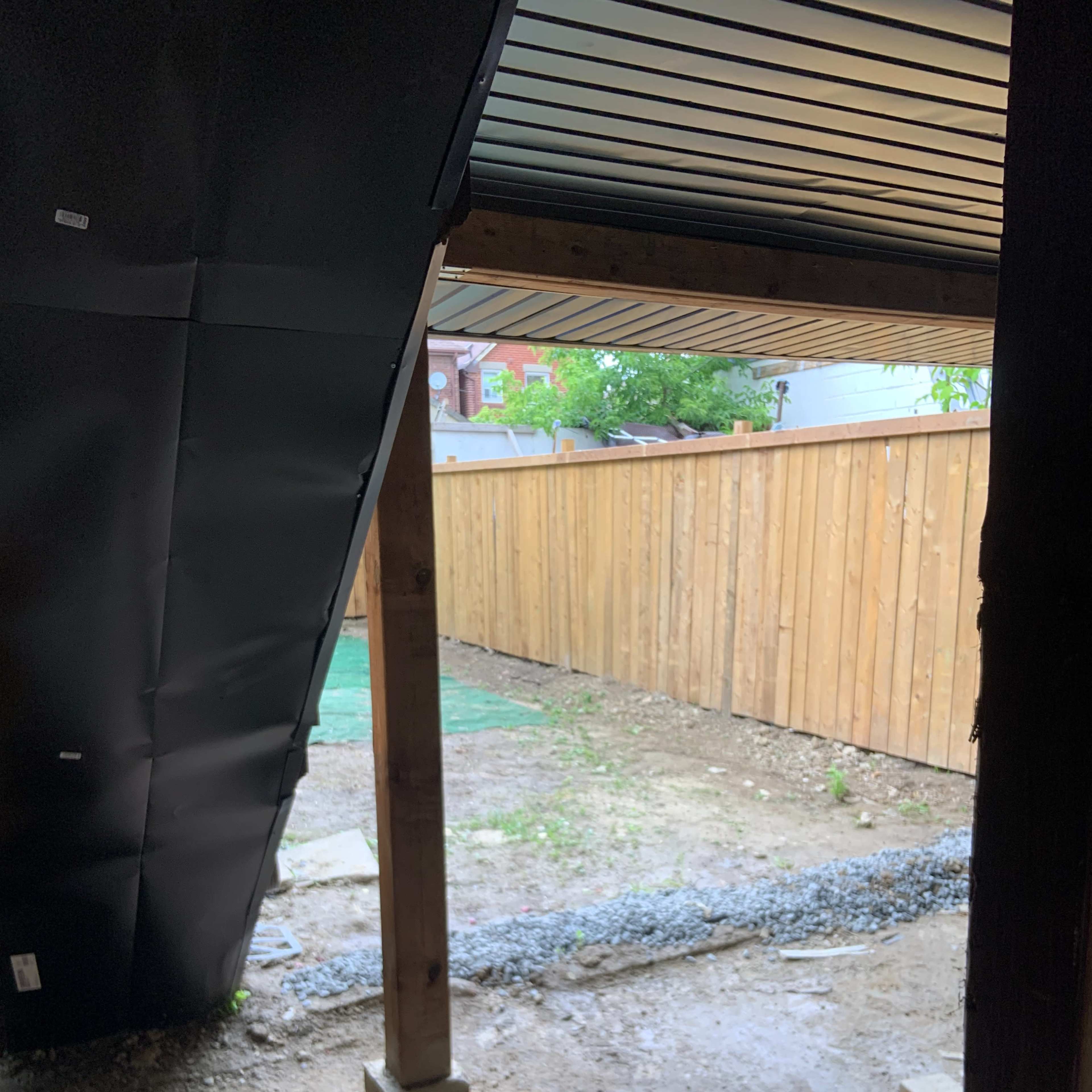 A covered area beneath a structure, revealing a gravel path leading to a fenced backyard space with grass and dirt.