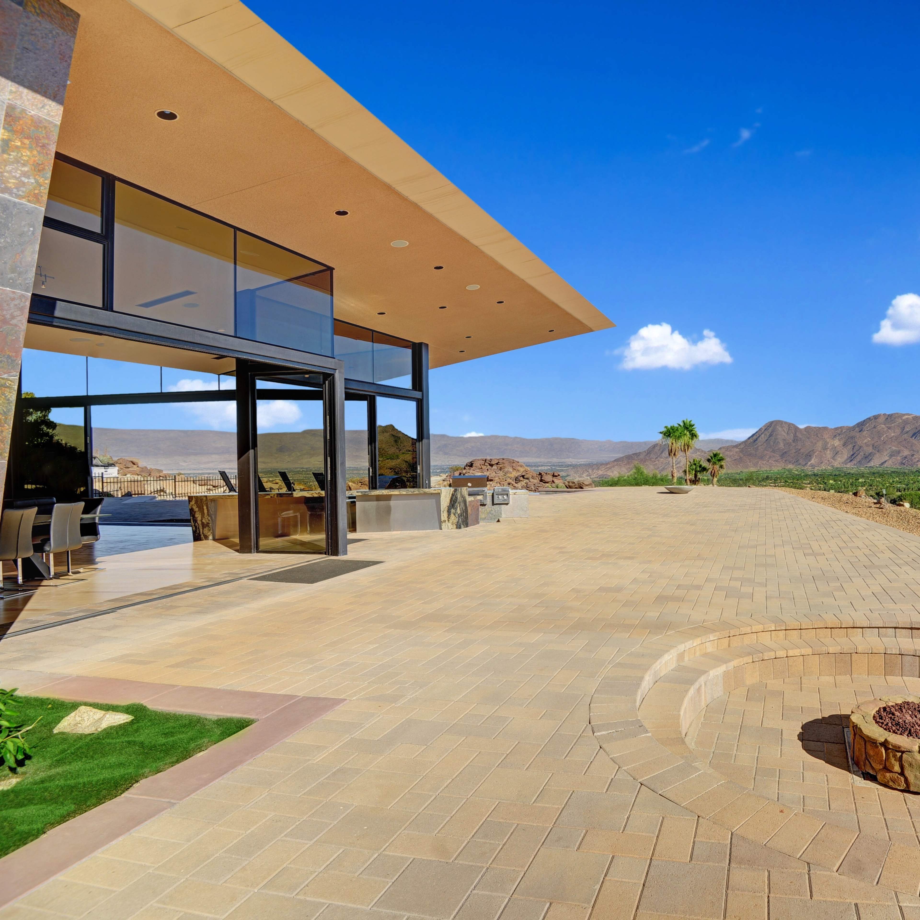 A modern building with large windows overlooks a spacious patio featuring a circular fire pit and a backdrop of mountains.