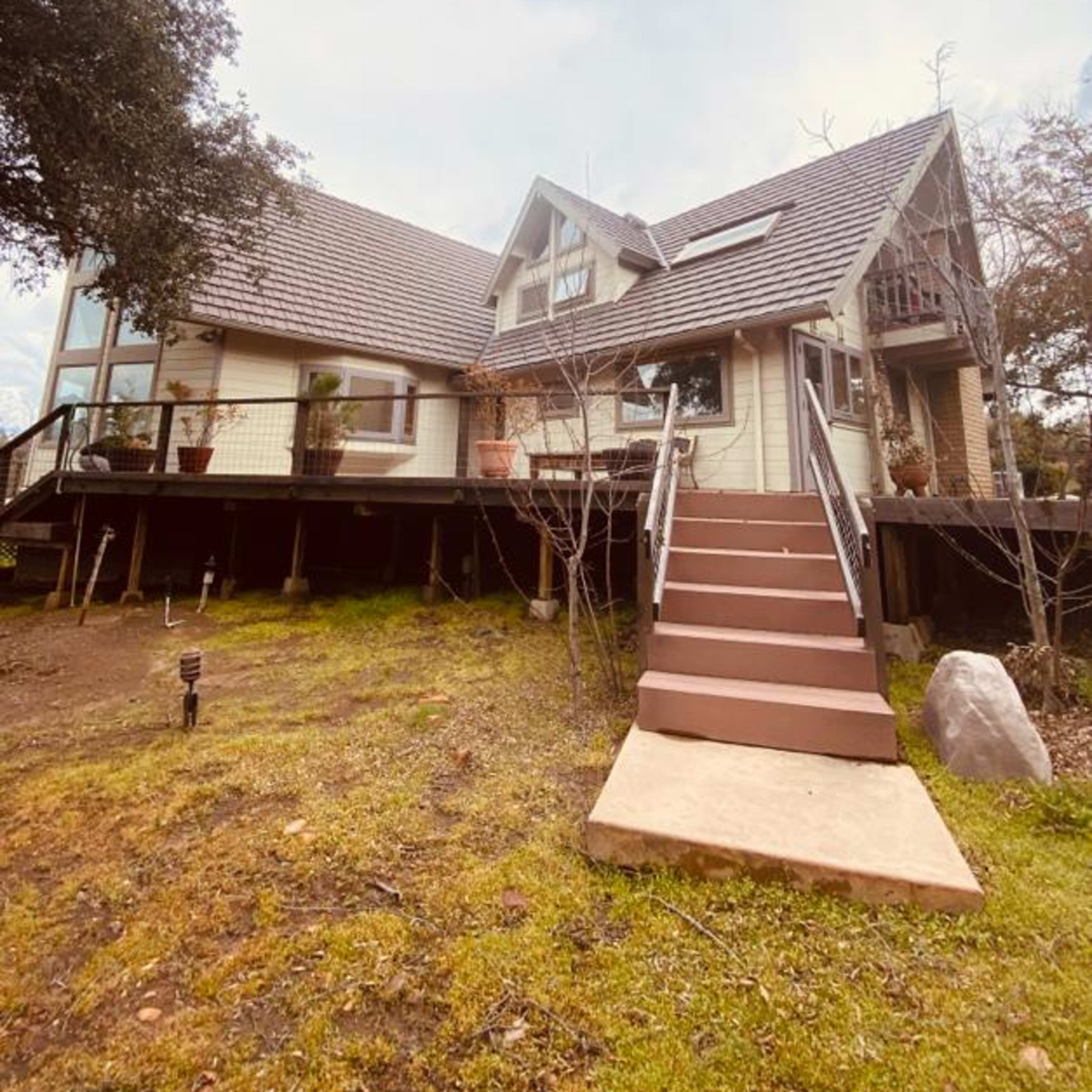 A two-story house with a steep roof and a front staircase is surrounded by grass and trees.