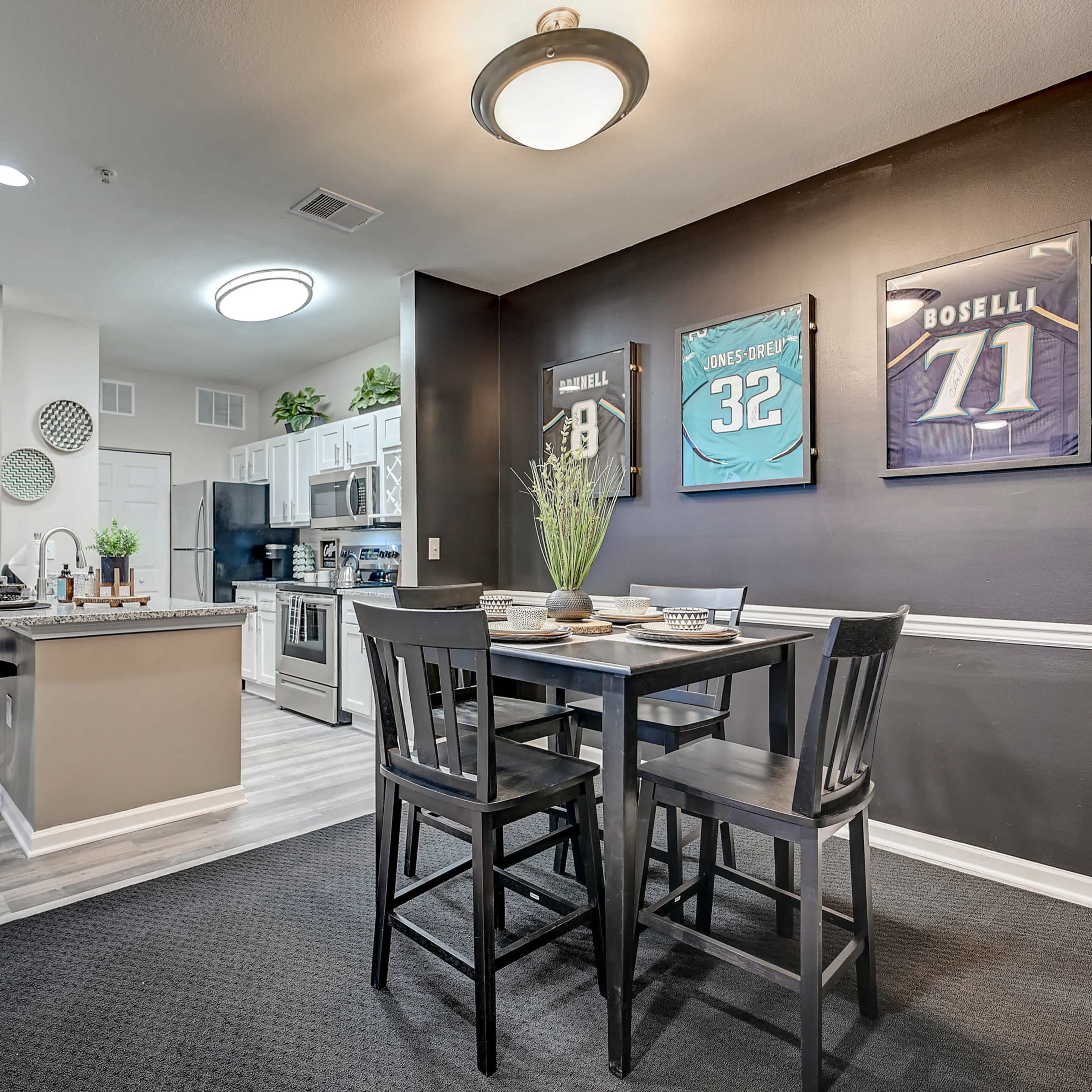 A modern kitchen and dining area featuring a black dining set, sports jerseys framed on the wall, and a well-equipped kitchen with white cabinets.