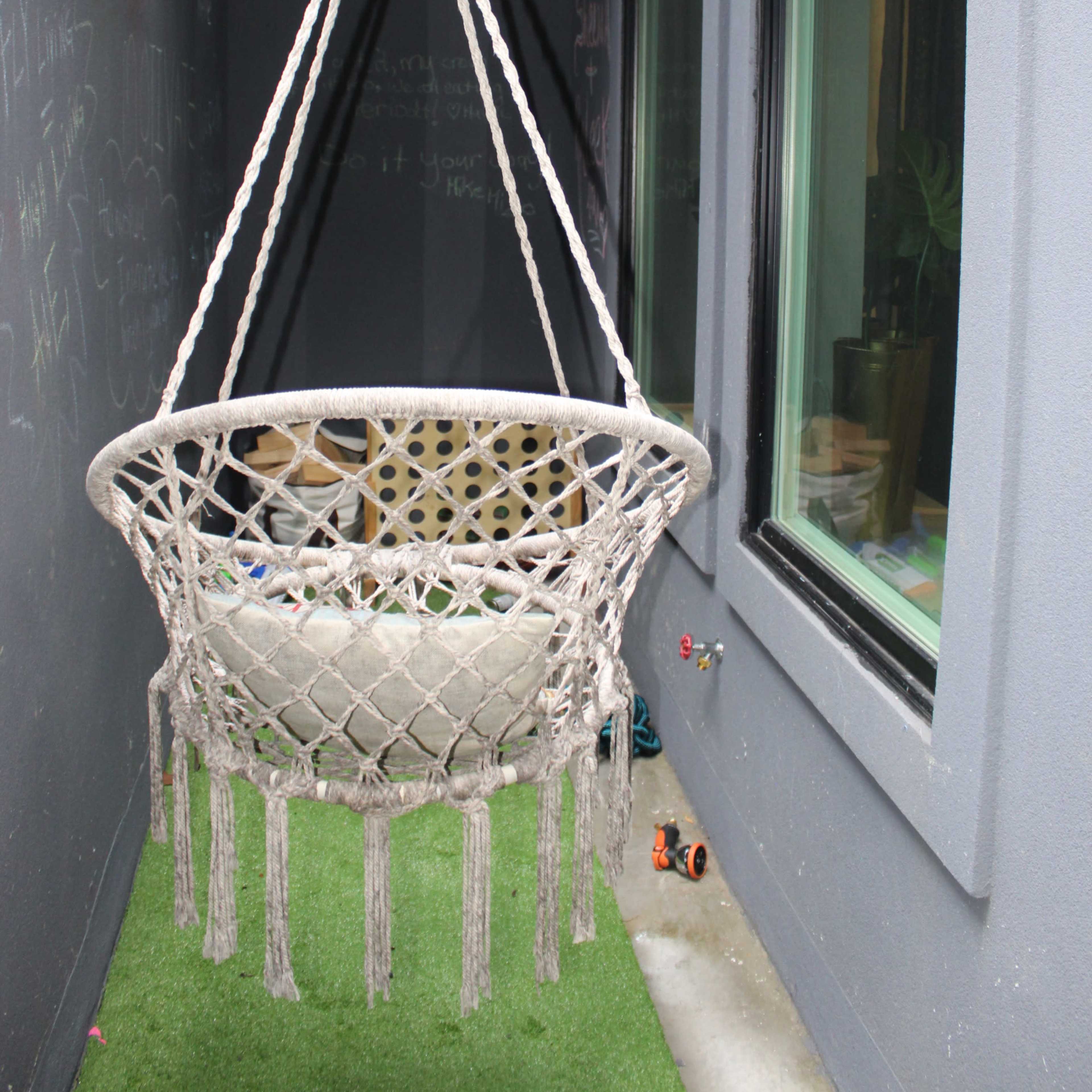 A macramé hanging chair is suspended in a narrow, covered space with green walls and artificial grass.