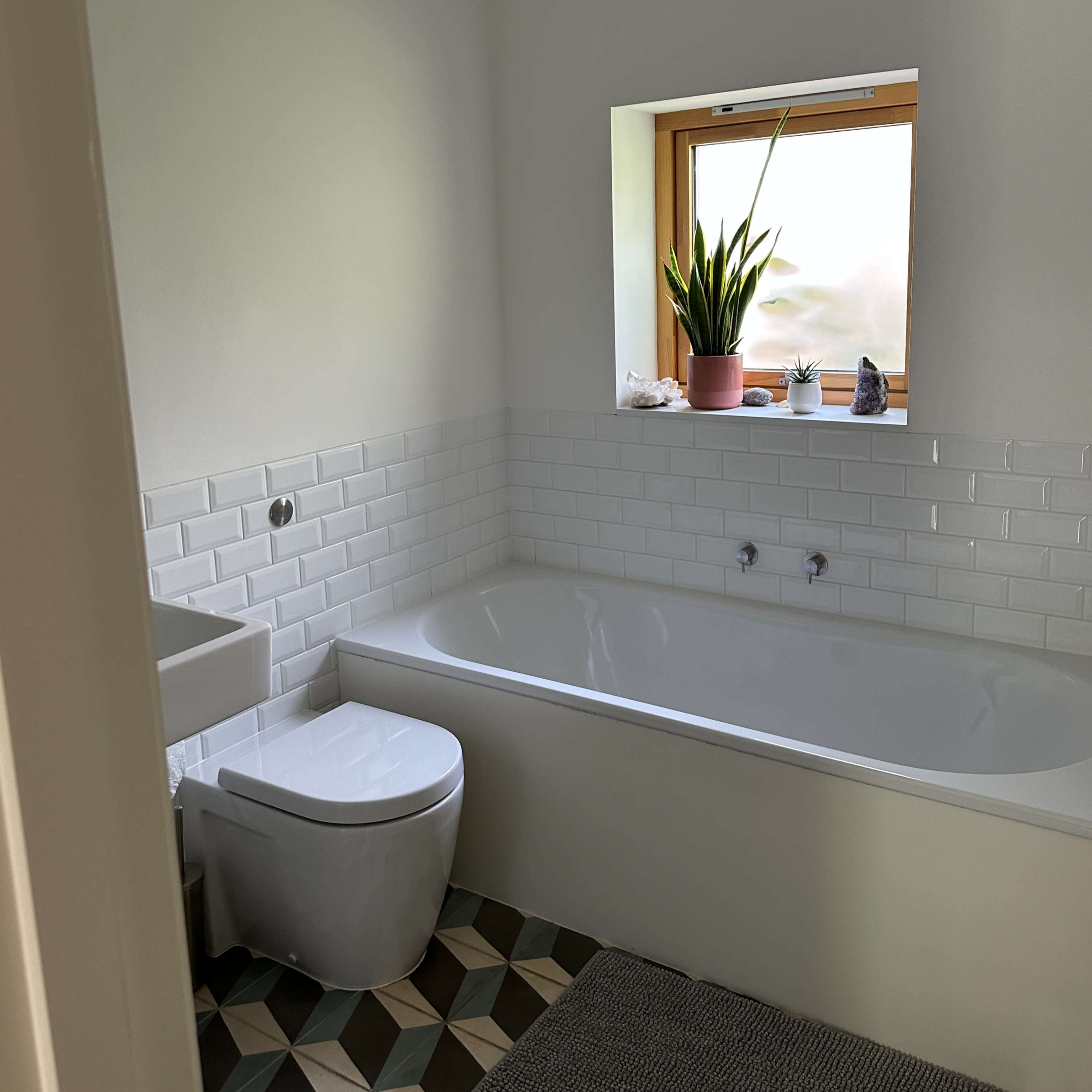 A bathroom features a white bathtub beside a wall of white tiles, a modern toilet, and a window with a potted plant.