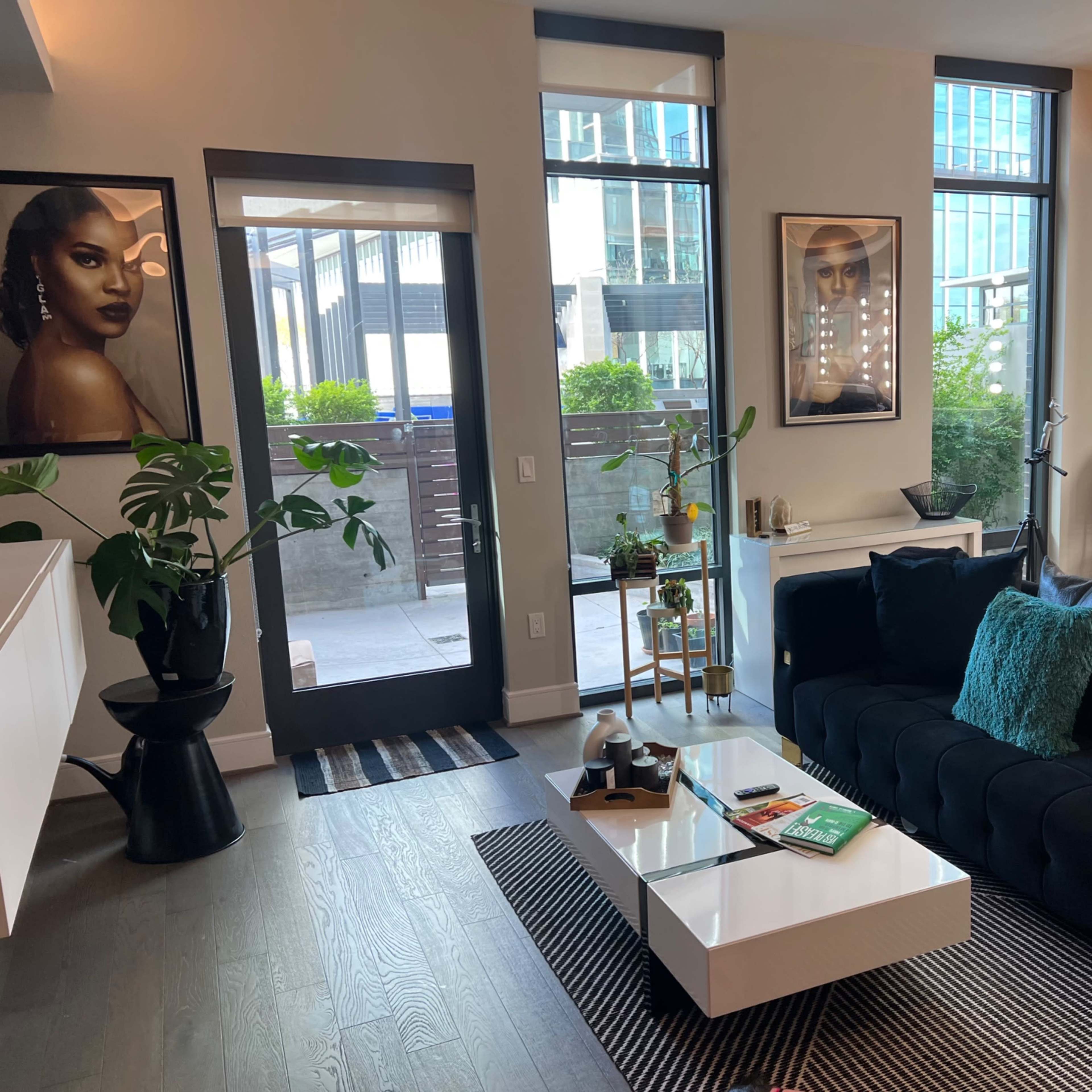 The scene features a modern living room with a black sofa, a coffee table, and large windows that let in natural light, along with decorative plants and art on the walls.