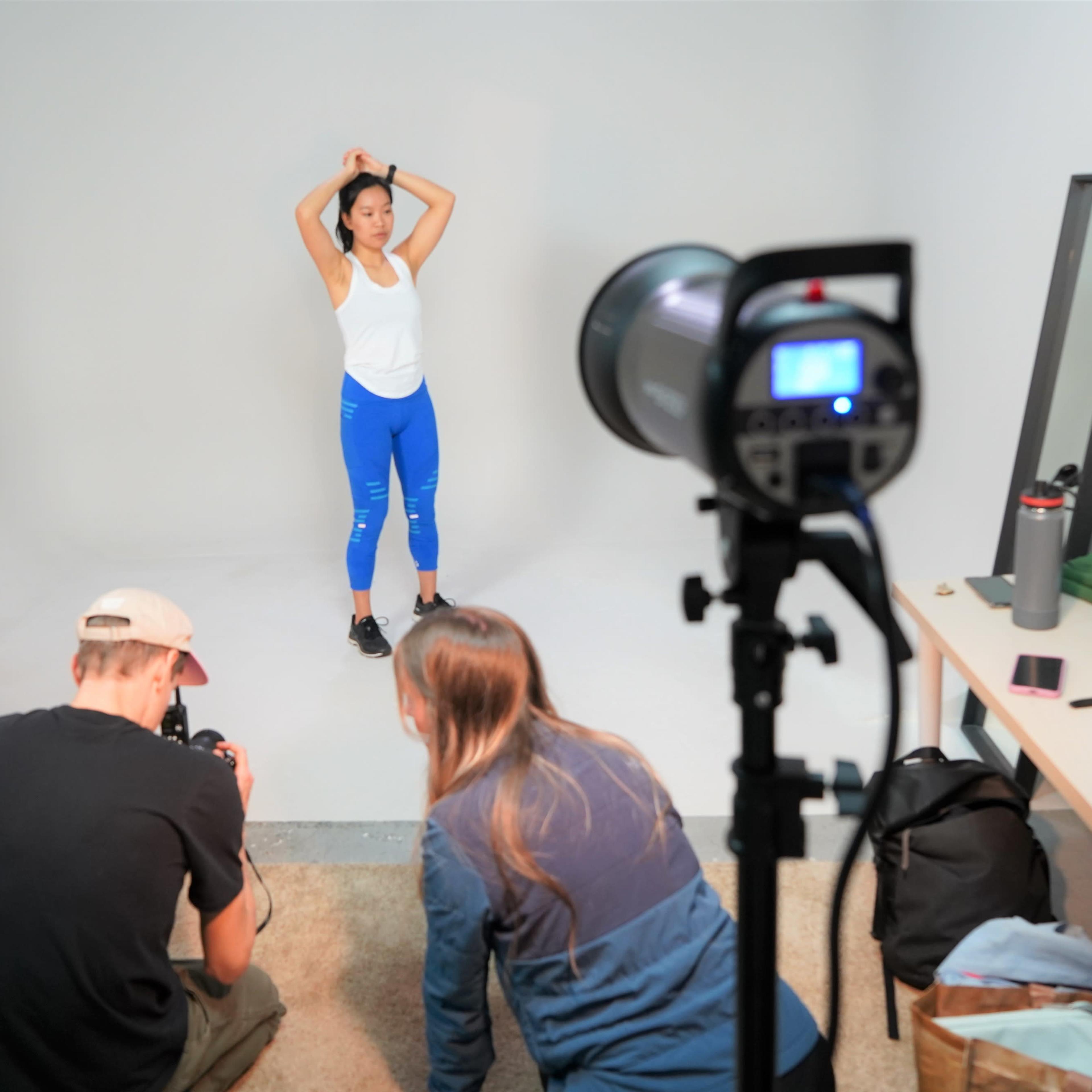 A model poses in a studio while two photographers set up equipment and capture images.