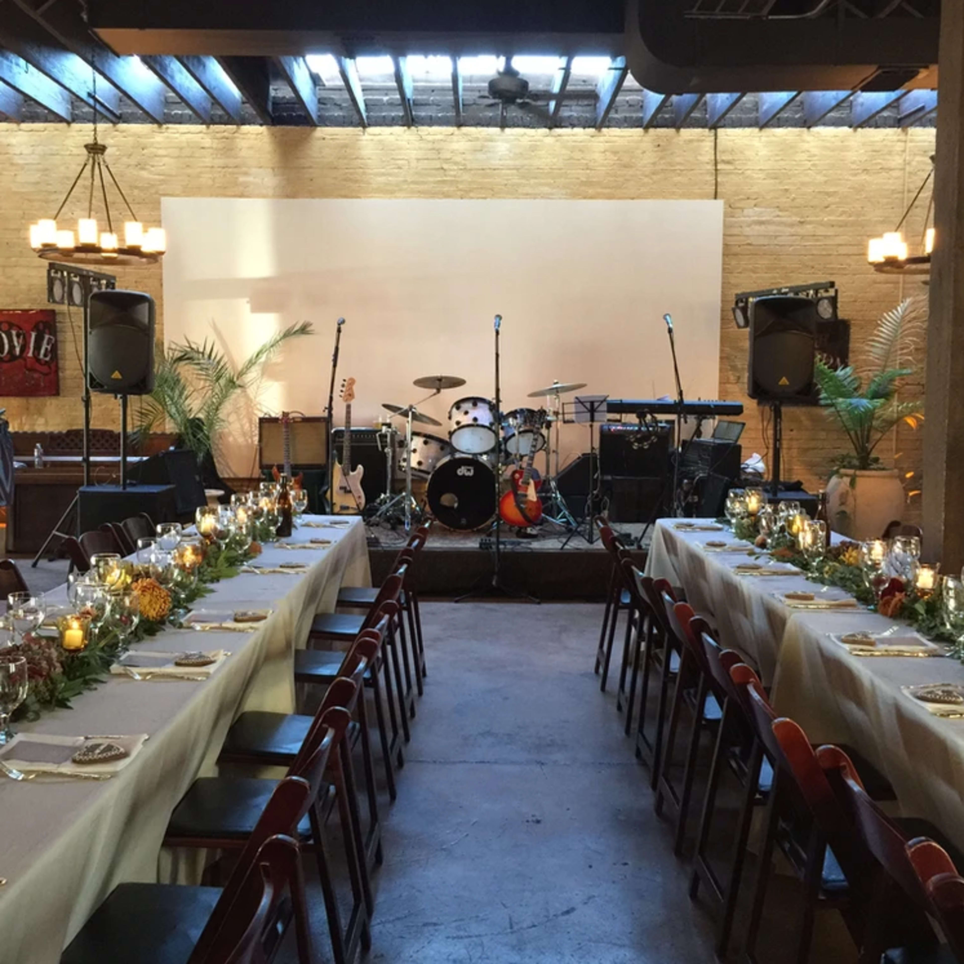 A spacious event venue is set up for a dinner with long tables adorned with greenery and candles, and a stage featuring musical instruments in the background.