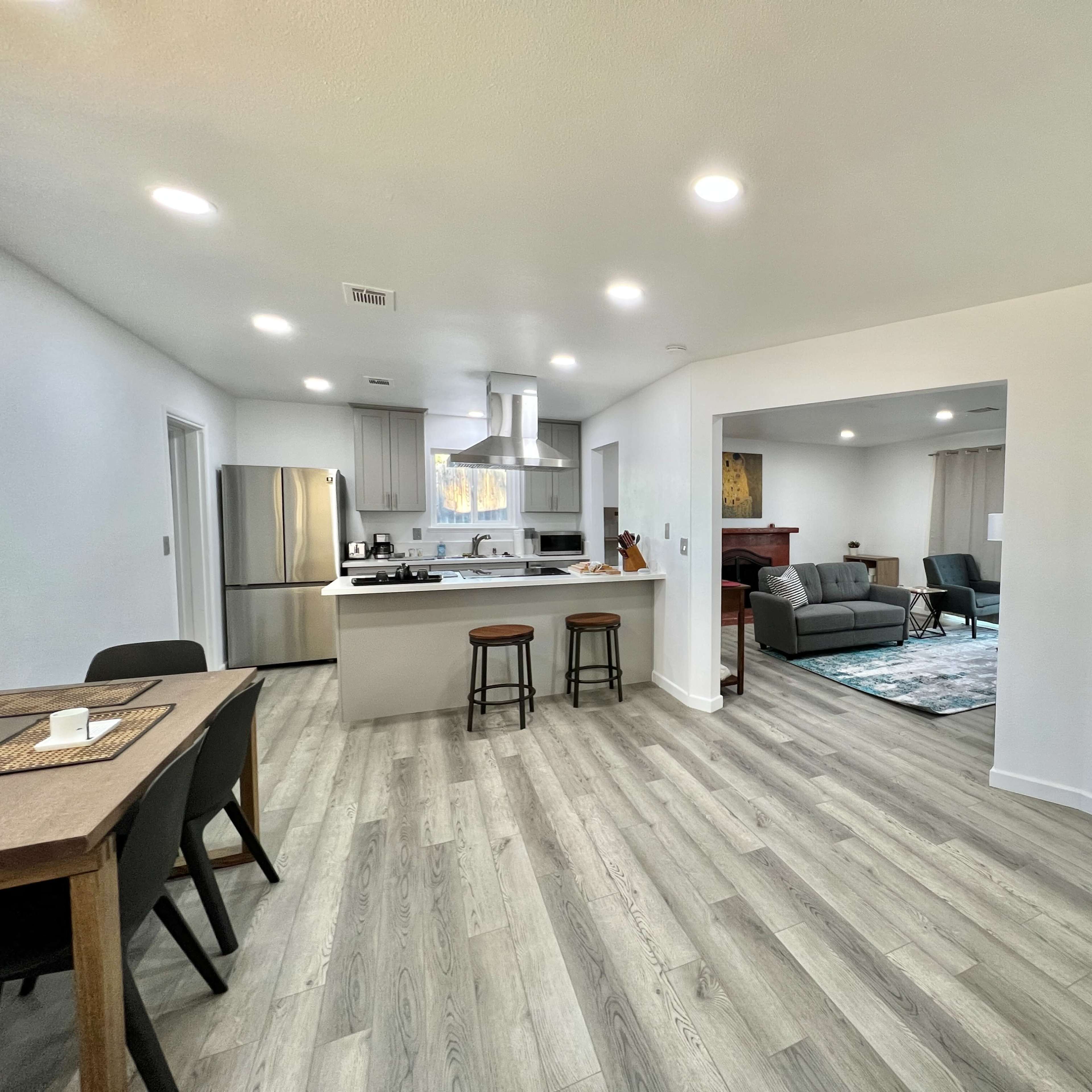 A modern, open-concept living space featuring a kitchen with stainless steel appliances, a dining area with two stools, and a view into a living room with a couch and a coffee table.