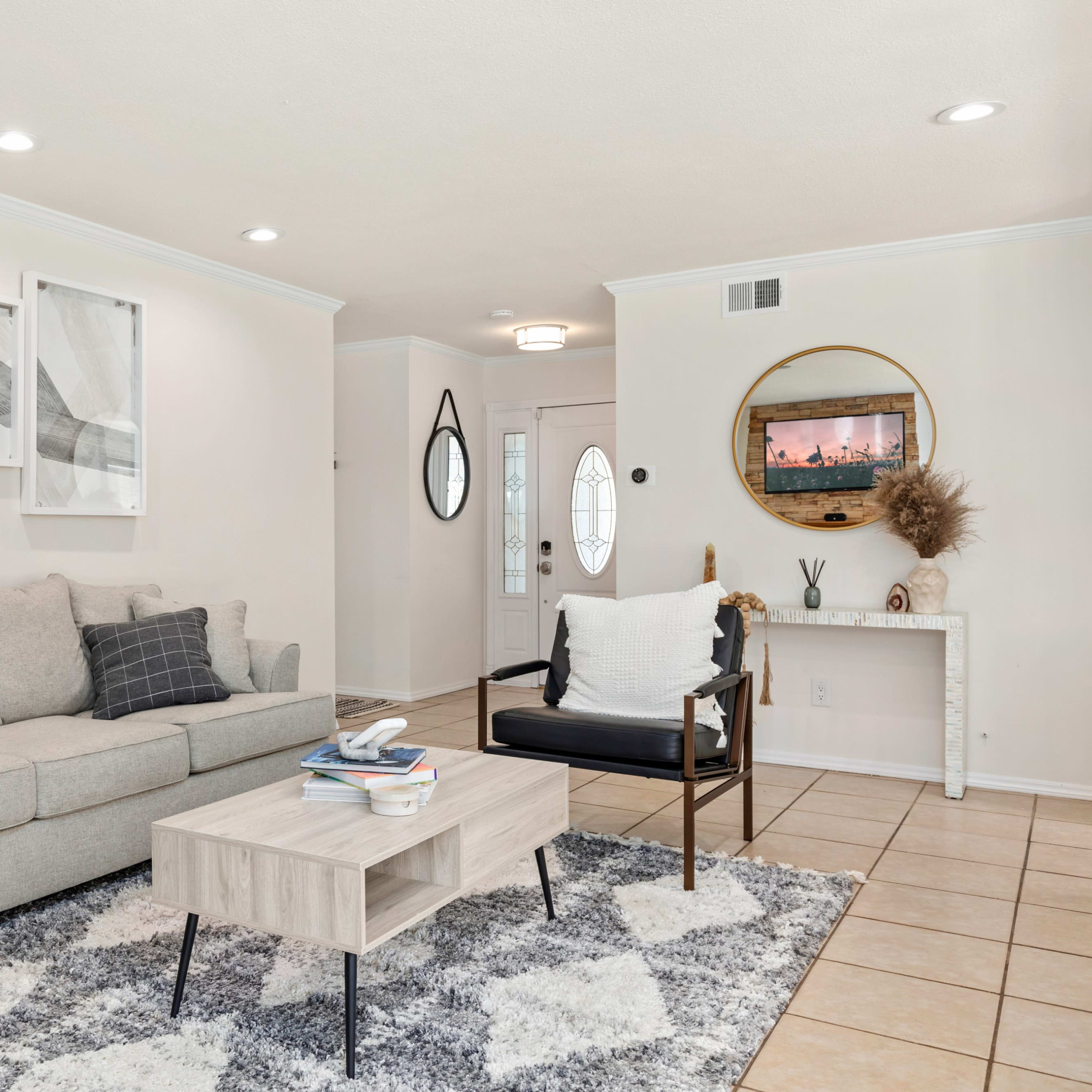A neutral-toned living room features a sofa with patterned cushions, a coffee table, and a side chair, all set against an entryway with a decorative mirror and a door leading outside.