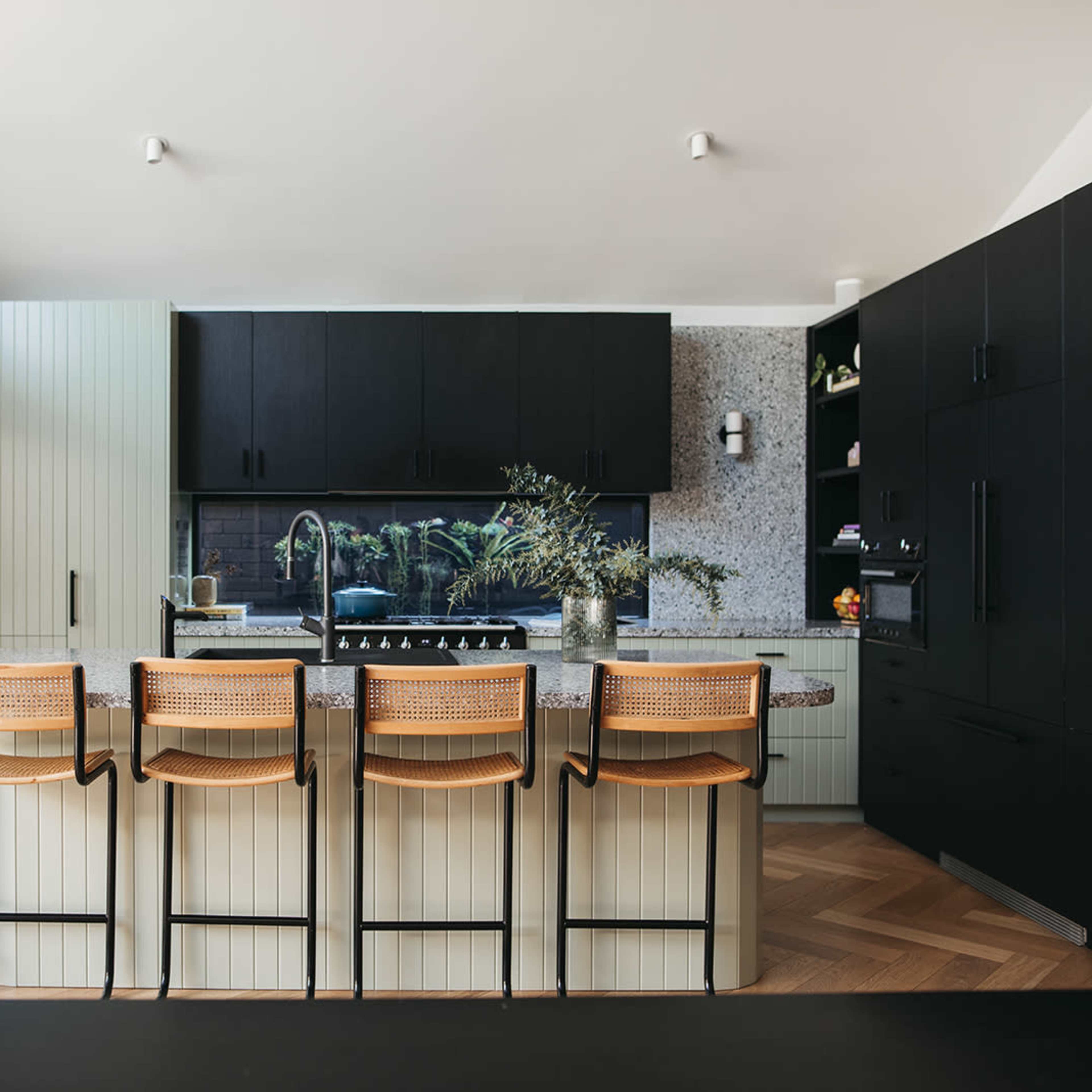 A modern kitchen features a large island with four woven bar stools, black cabinetry, and a light green pantry against a textured wall.