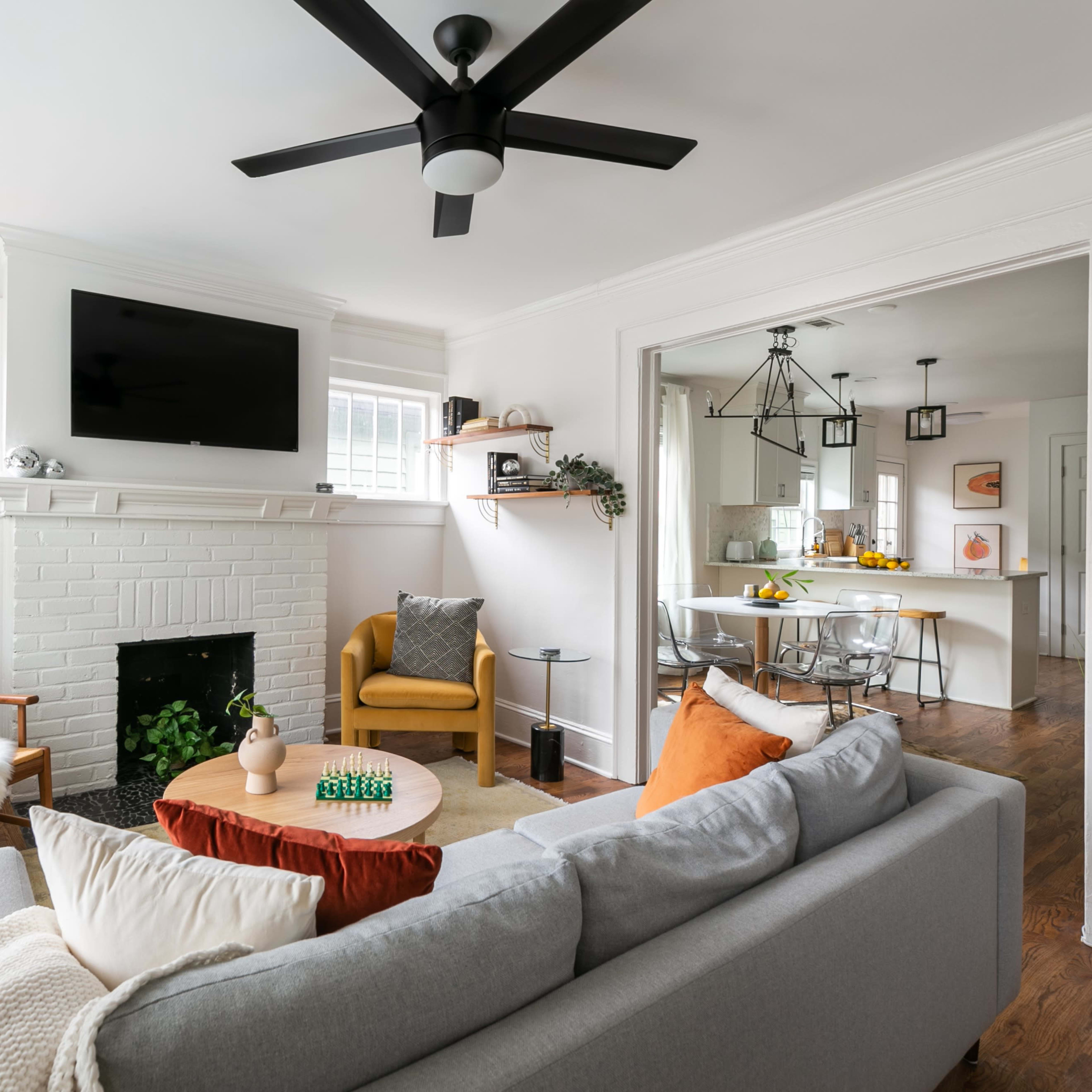 A modern living room features a gray sofa, a coffee table, and a white brick fireplace with a flat-screen TV mounted above it, leading into a bright dining area.