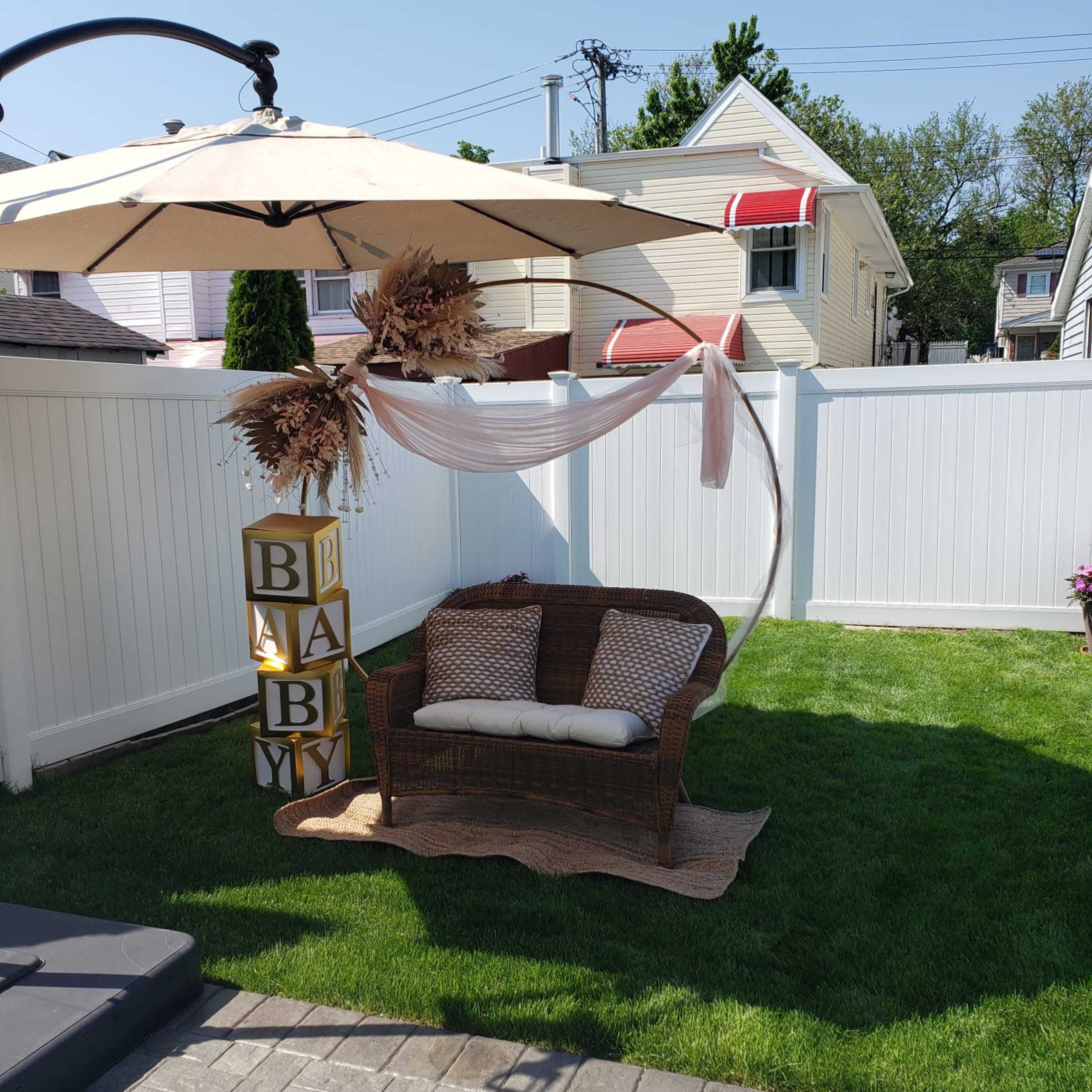 A wicker loveseat is positioned under a large umbrella, accompanied by decorative blocks spelling "BABY" and surrounded by a neatly maintained grass lawn and white fence.