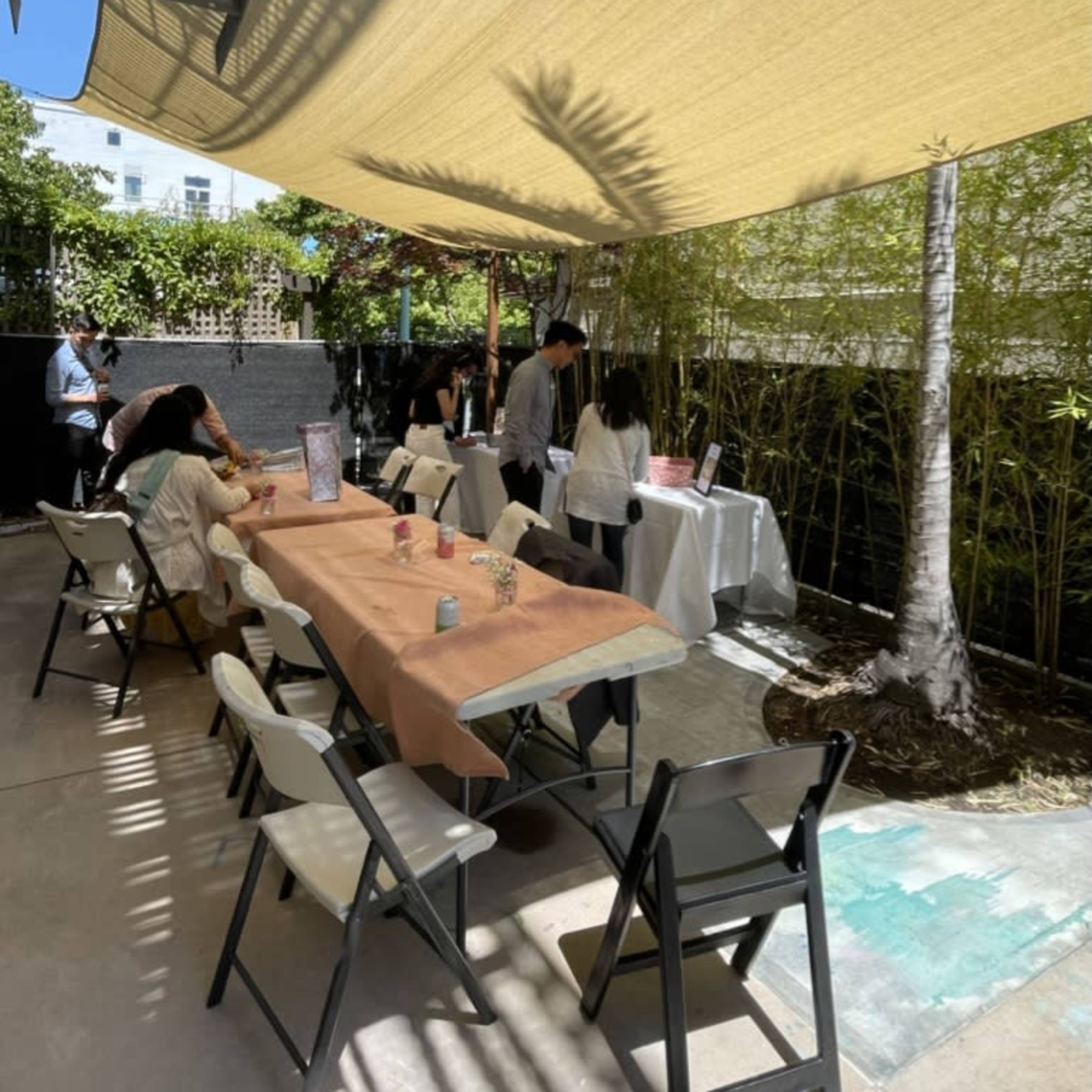 A shaded outdoor area features tables with patrons, surrounded by greenery and sunlight filtering through a canopy.