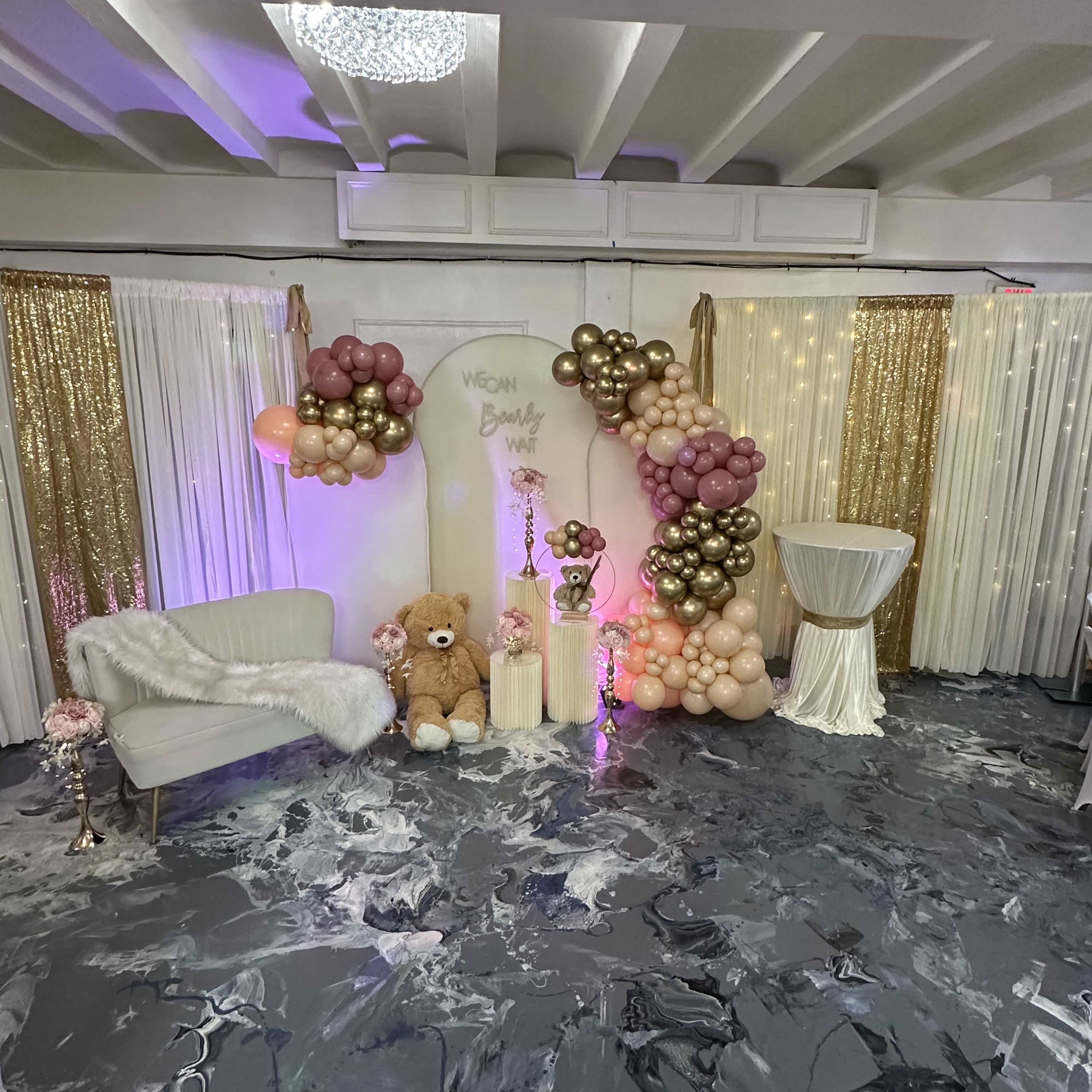 An event decor setup featuring a balloon arch, a teddy bear, a small table, and backdrop curtains adorned with lights.