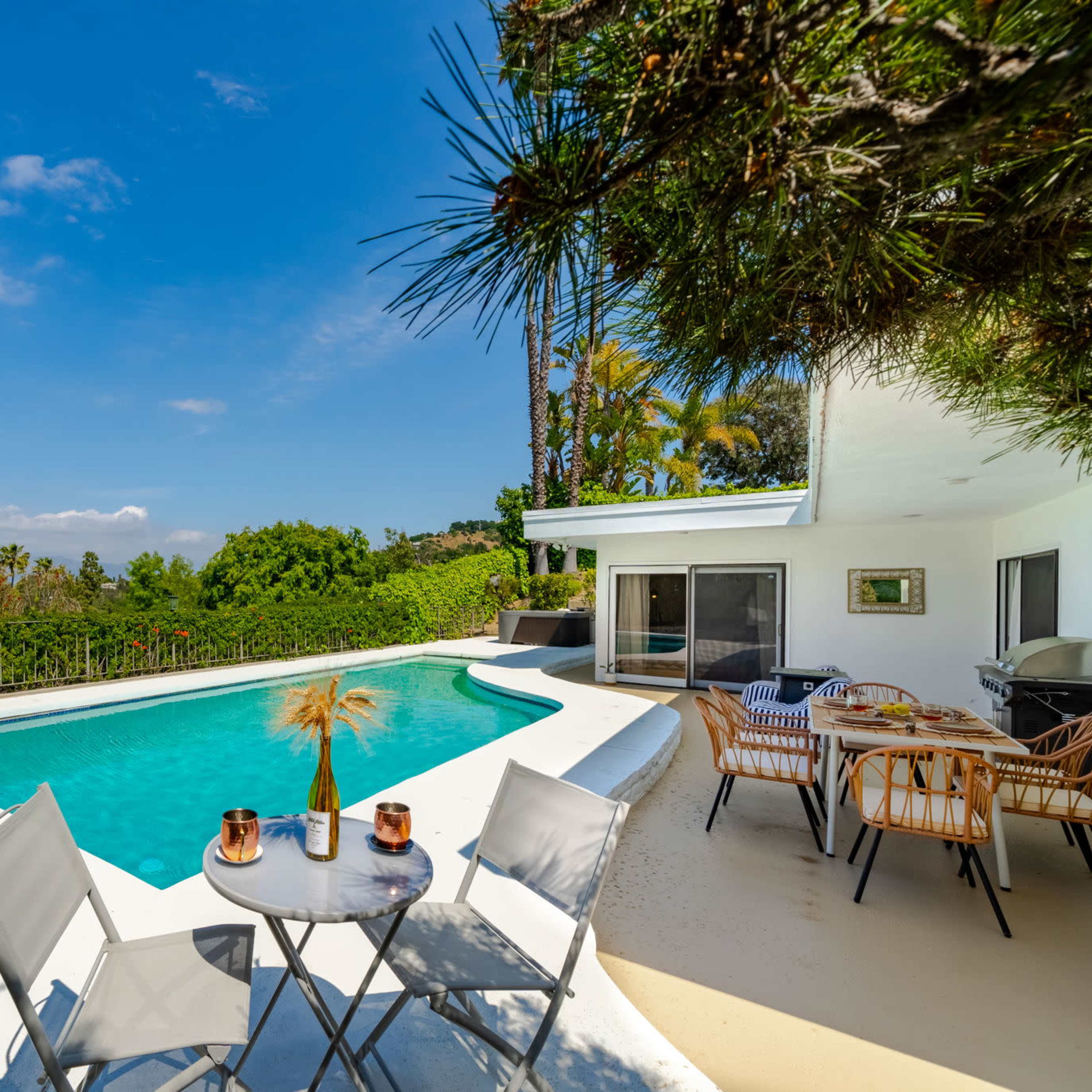 A modern outdoor area featuring a swimming pool, a small table with chairs, and a patio that overlooks a lush landscape.