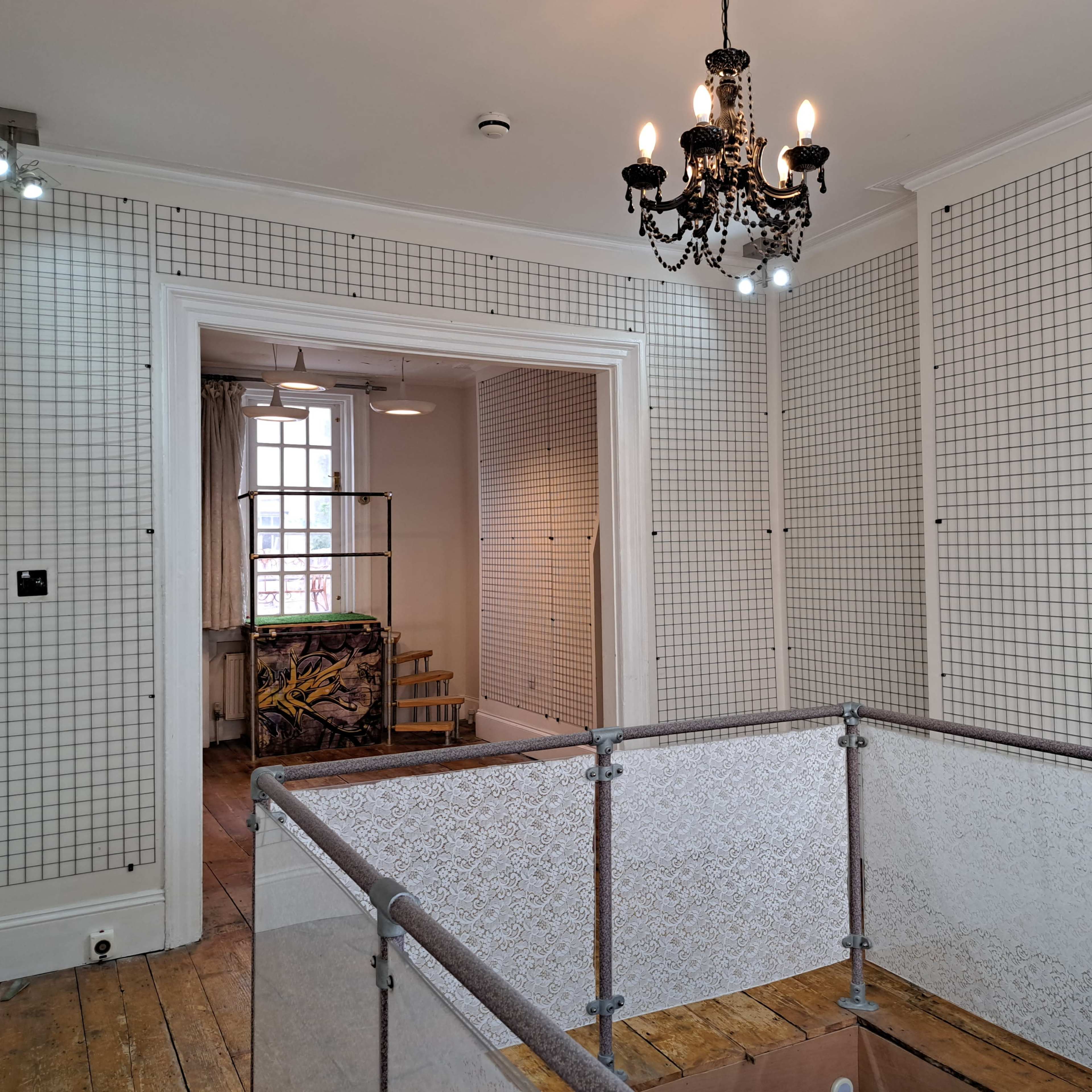 A room with grid-patterned walls, a chandelier, and a railing overlooking a partially visible space with wooden steps and a cluttered area behind a doorway.