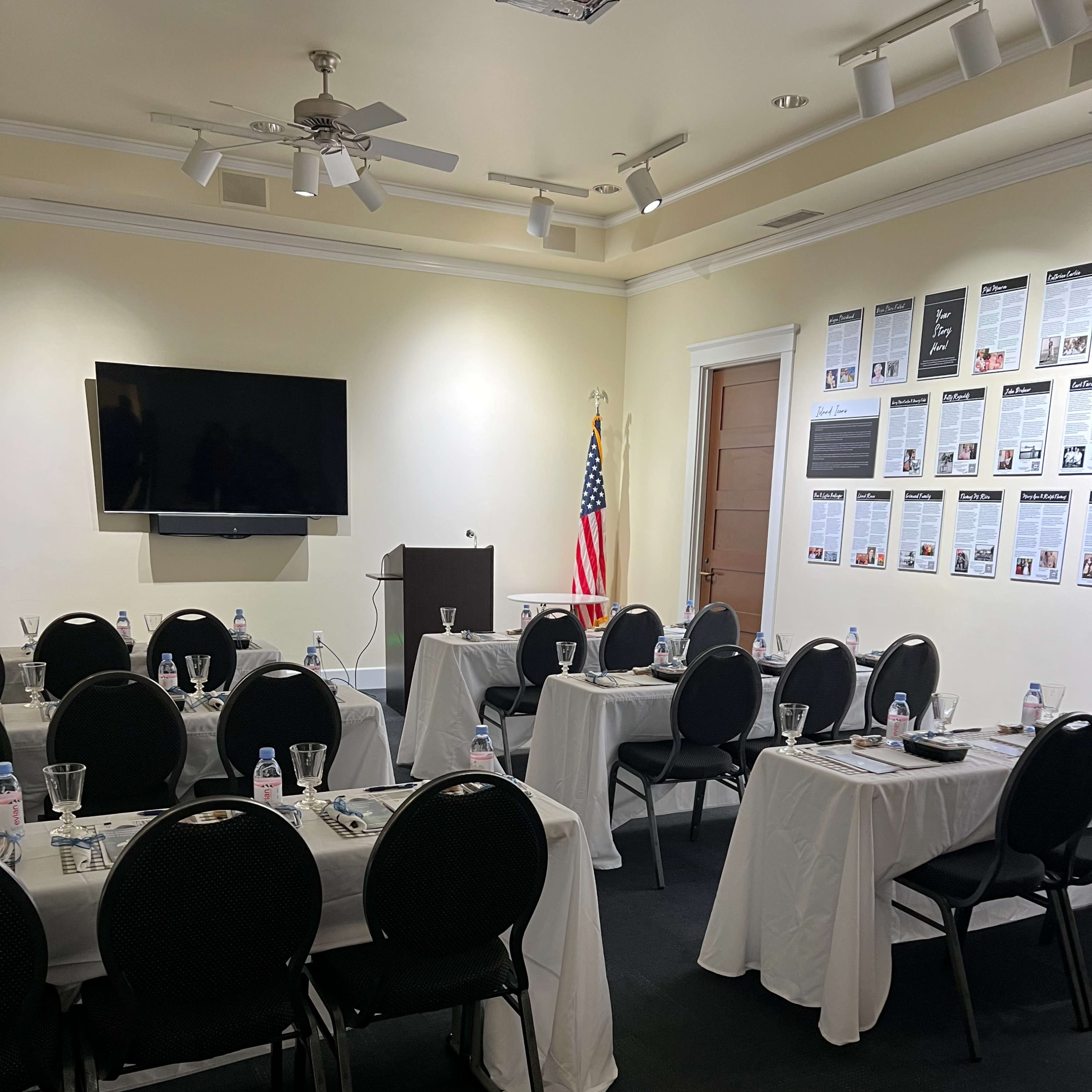 A conference room is set up with several tables covered in white linens, each arranged with water glasses and paper, along with a podium and an American flag in the background.
