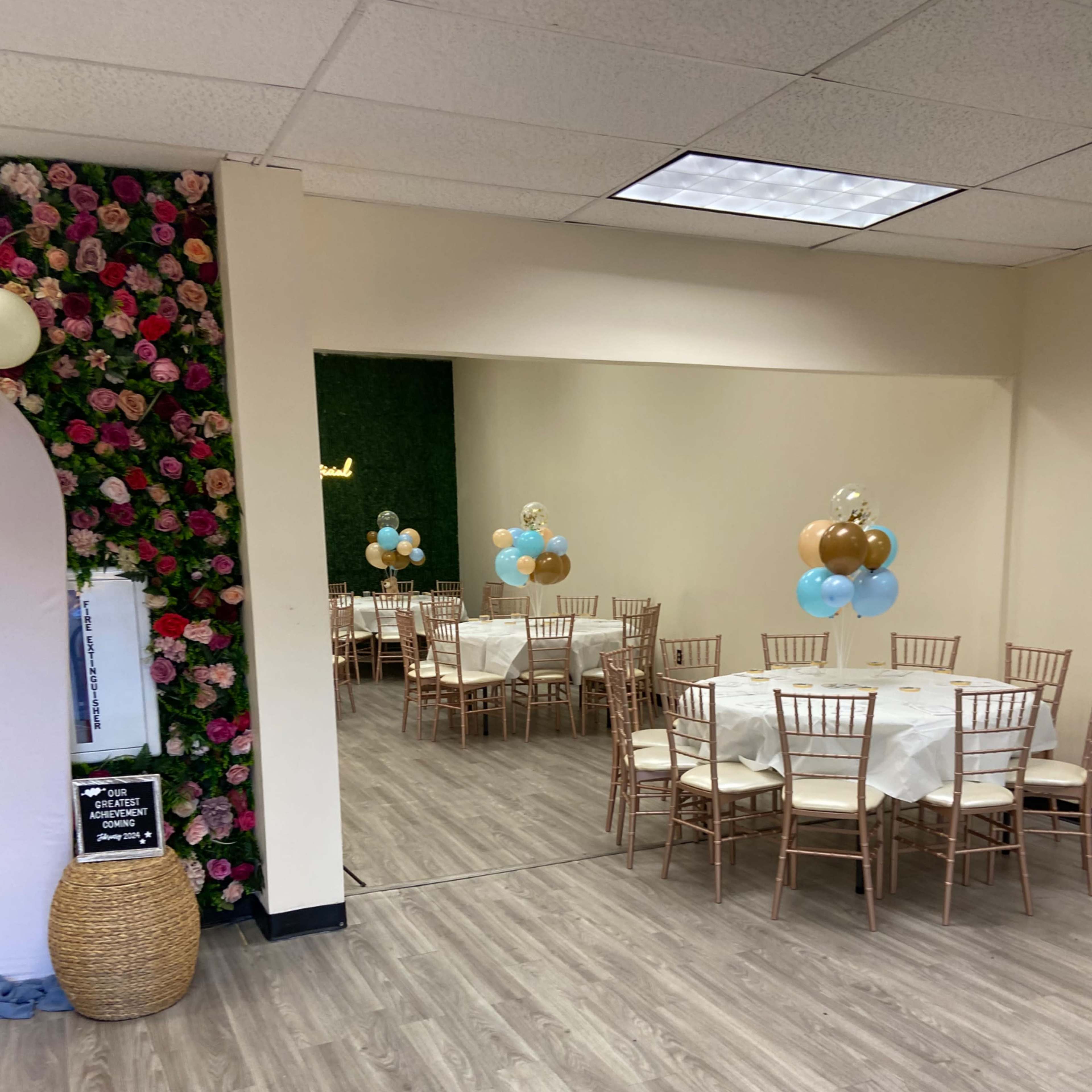 A decorated event space with wooden tables and chairs, a floral wall, and a mirrored area featuring balloons in blue and gold colors.