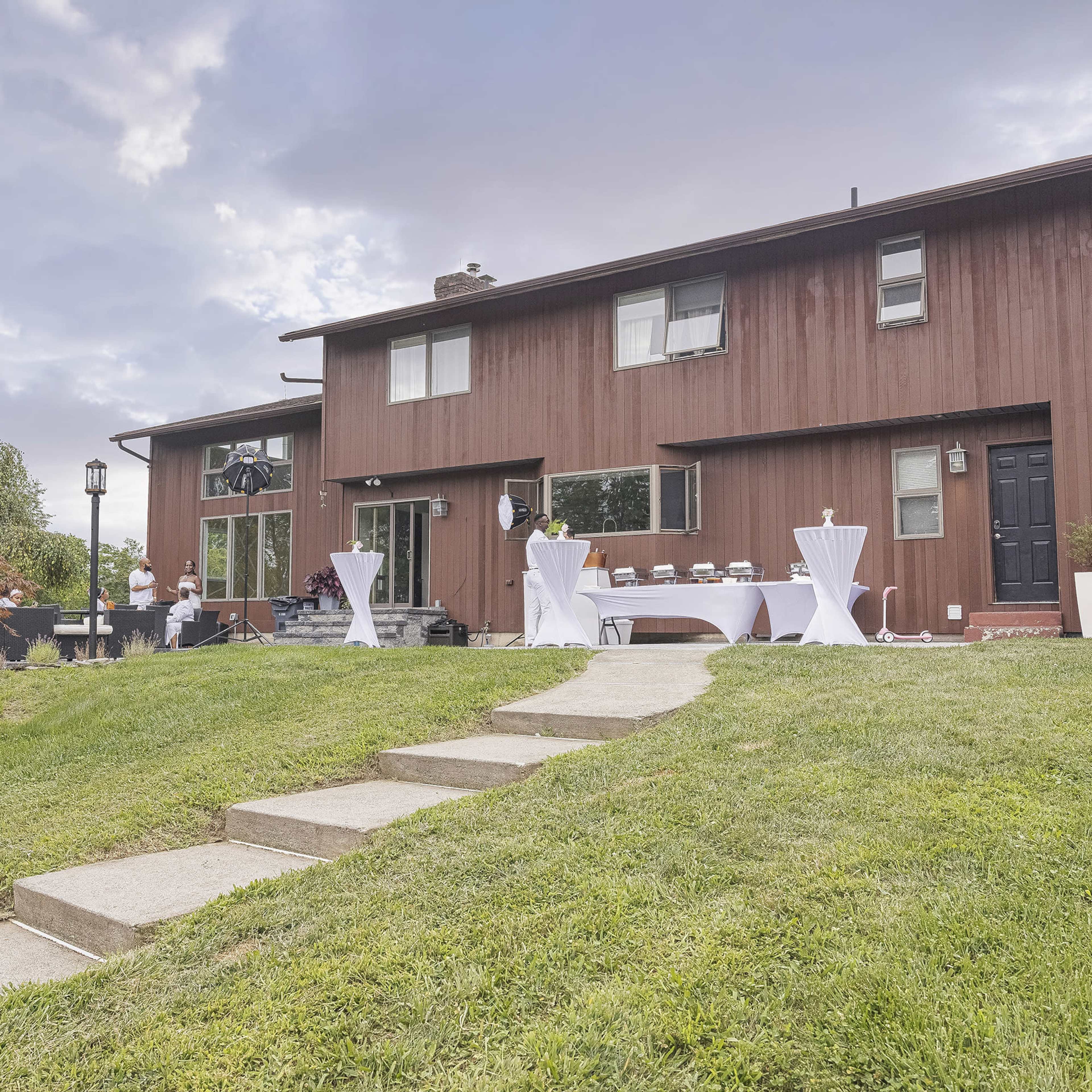 A modern brown house with a manicured lawn features several tables with white tablecloths arranged outside, and a few people are preparing for an event.
